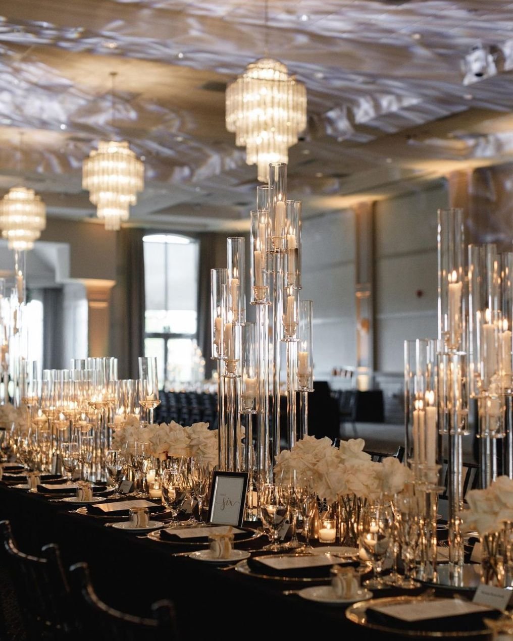 Elegant banquet table decorated with tall glass candle holders, white floral centerpieces, and gold tableware in a luxurious venue with chandeliers and large windows.