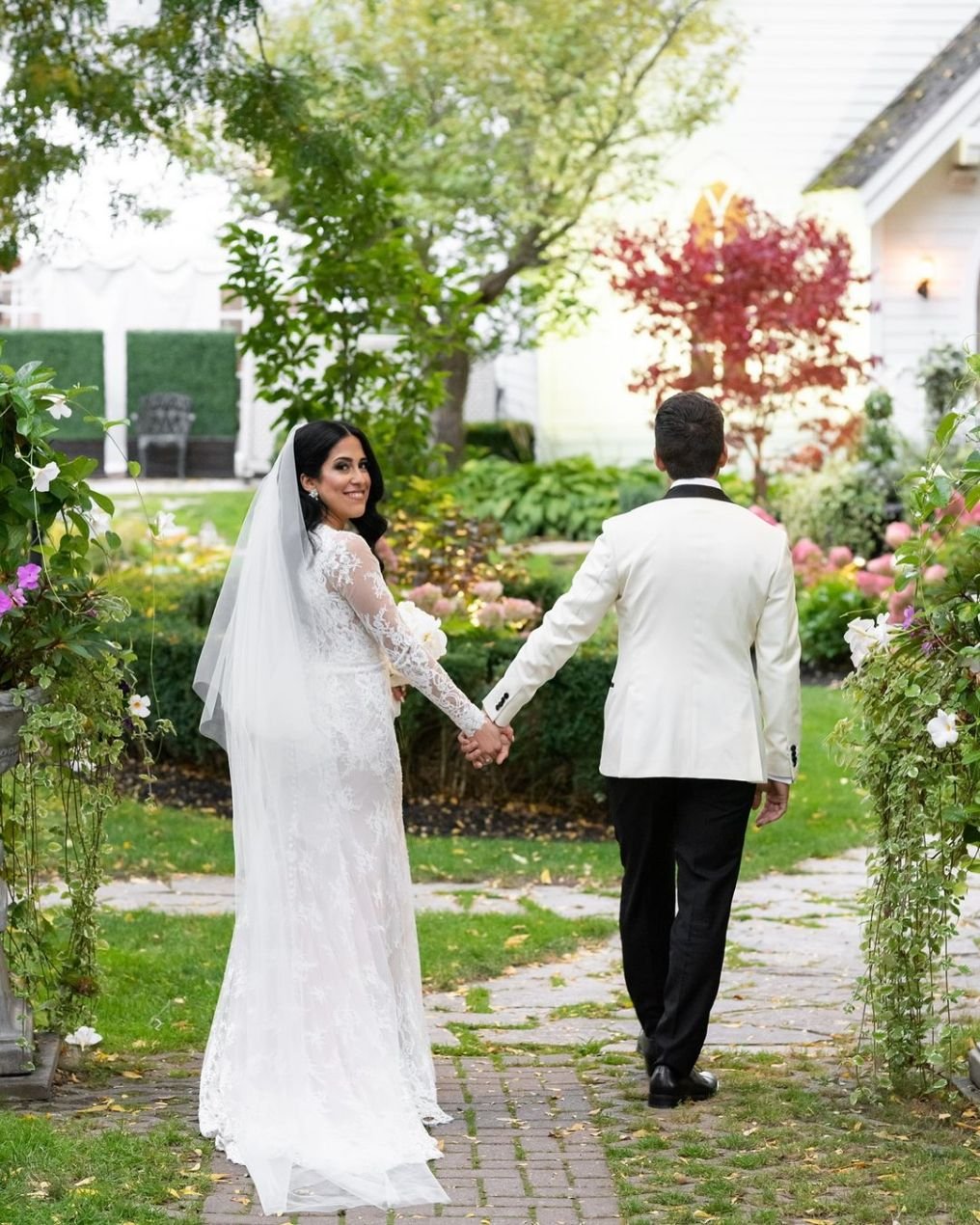 A bride and groom holding hands and walking in a garden, with the bride smiling and looking back at the camera.