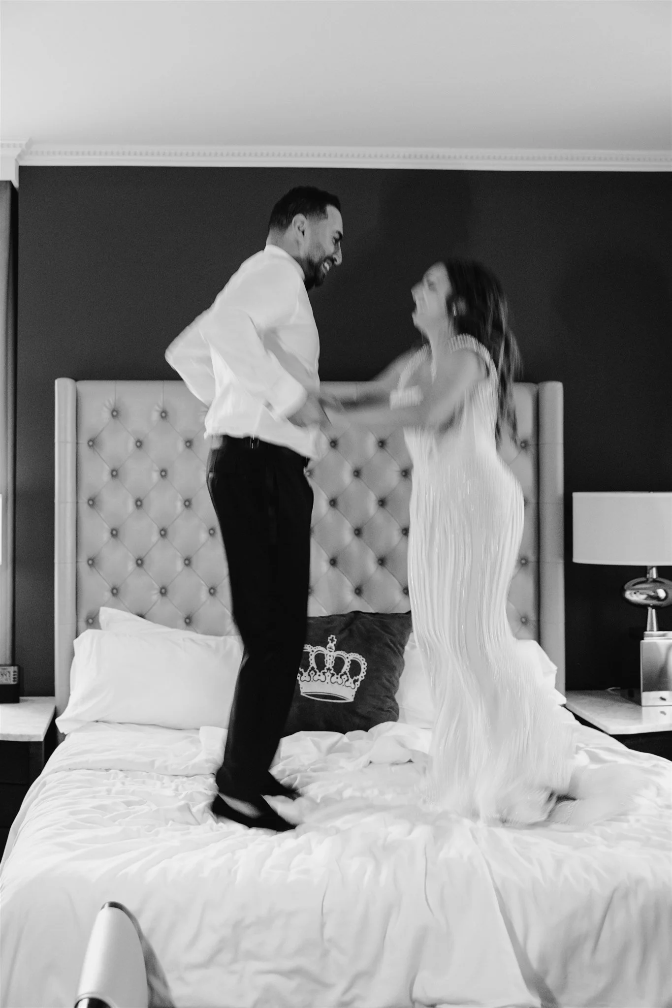 A man and a woman are on a bed, dancing and laughing together. The man is wearing a white shirt and dark pants, and the woman is wearing a long, flowing dress. There are pillows and bedside tables with lamps on each side of the bed.