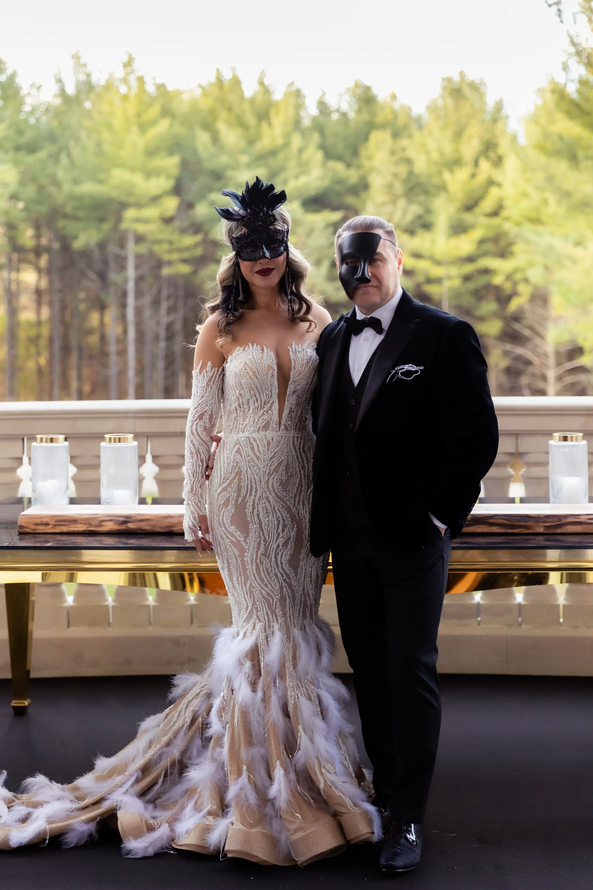 A bride and groom in formal attire wearing black masquerade masks, standing outdoors on a terrace with a forest in the background. The bride is in an elegant, feathered wedding gown, and the groom is in a black tuxedo.
