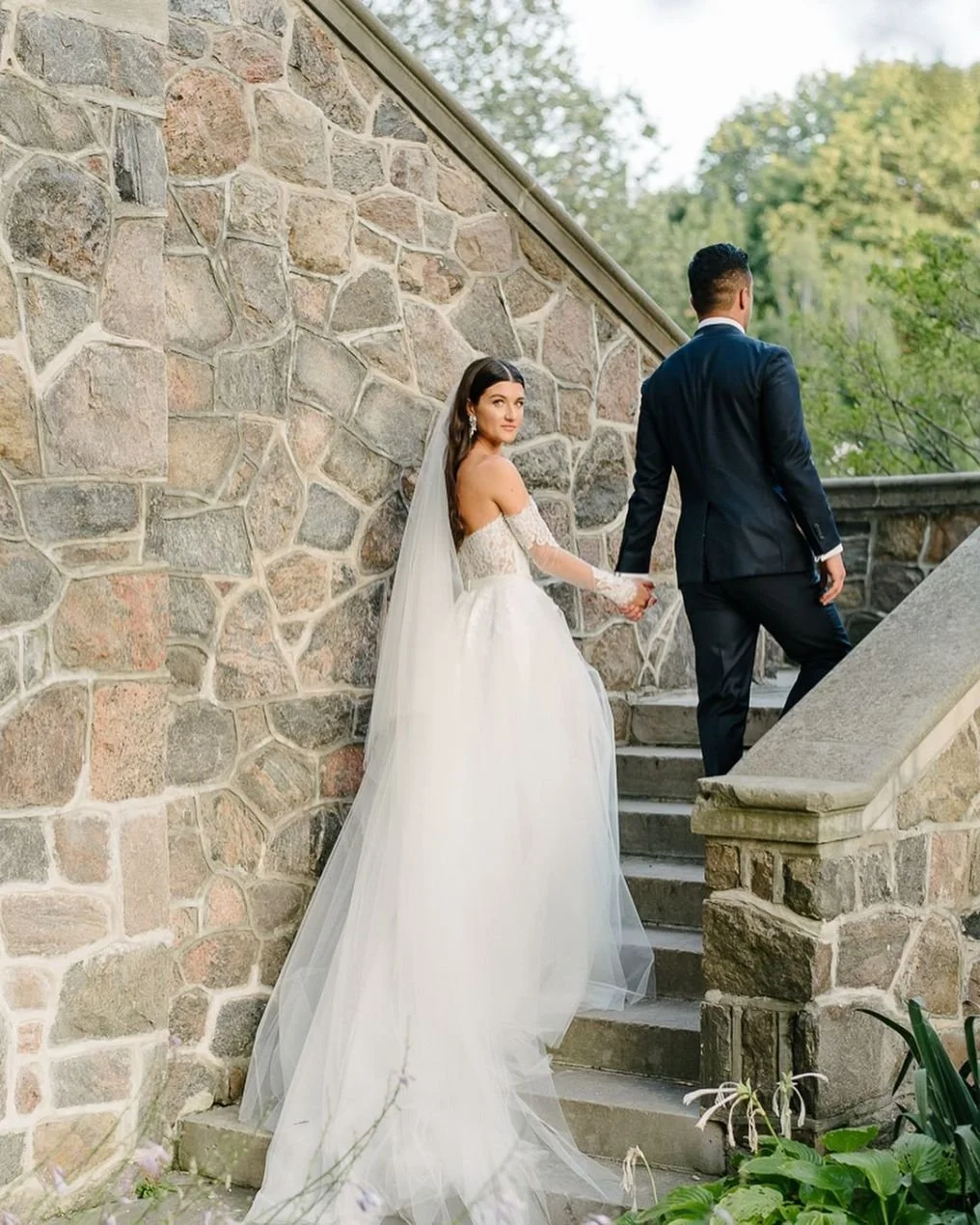 Bride and groom holding hands on outdoor stone staircase, with bride in a white wedding gown and veil looking back at the camera, groom in a dark suit walking ahead.