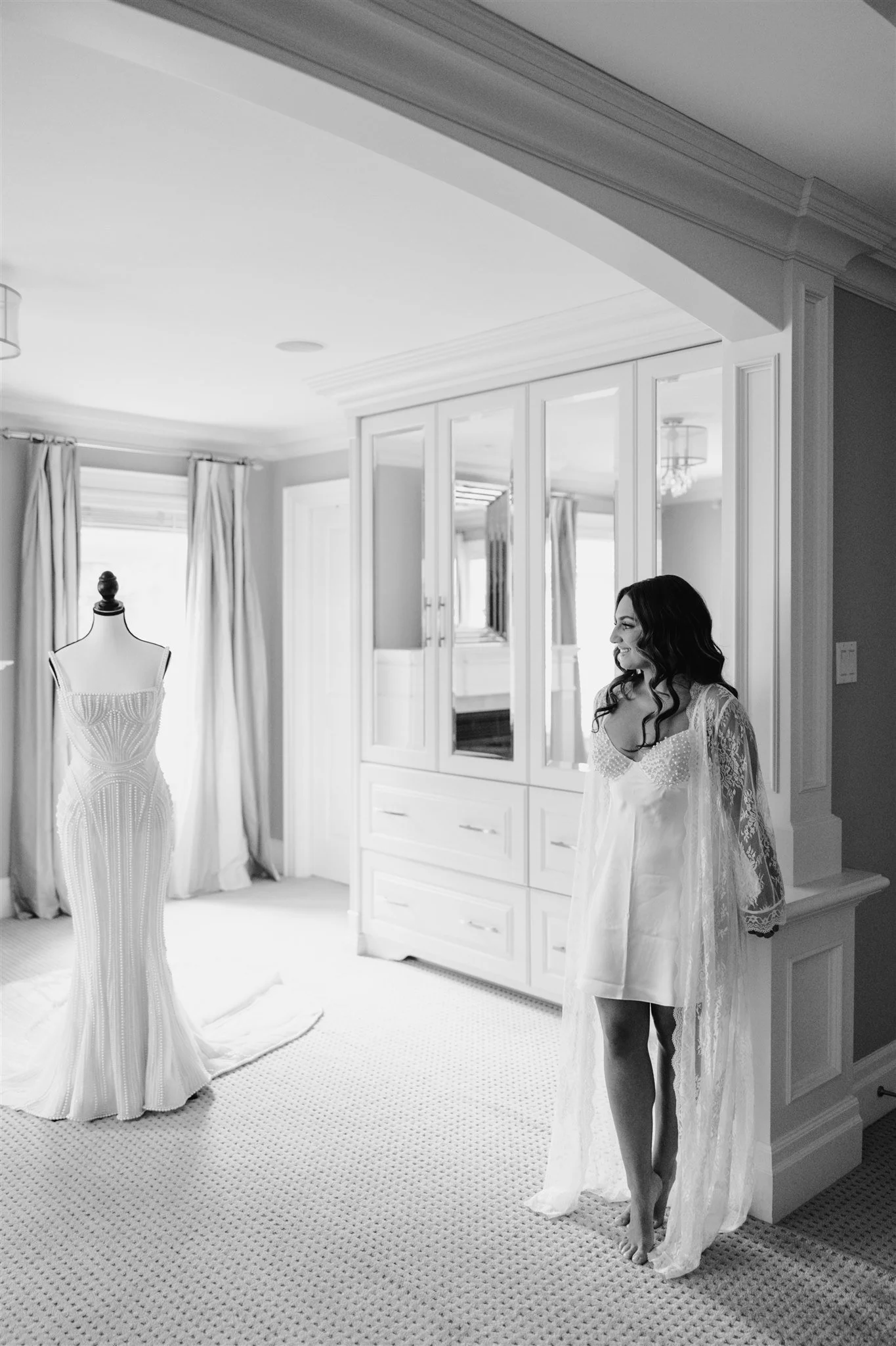 A woman in a lace robe stands in a well-lit room, looking at a wedding gown on a mannequin, with large windows and curtains in the background.