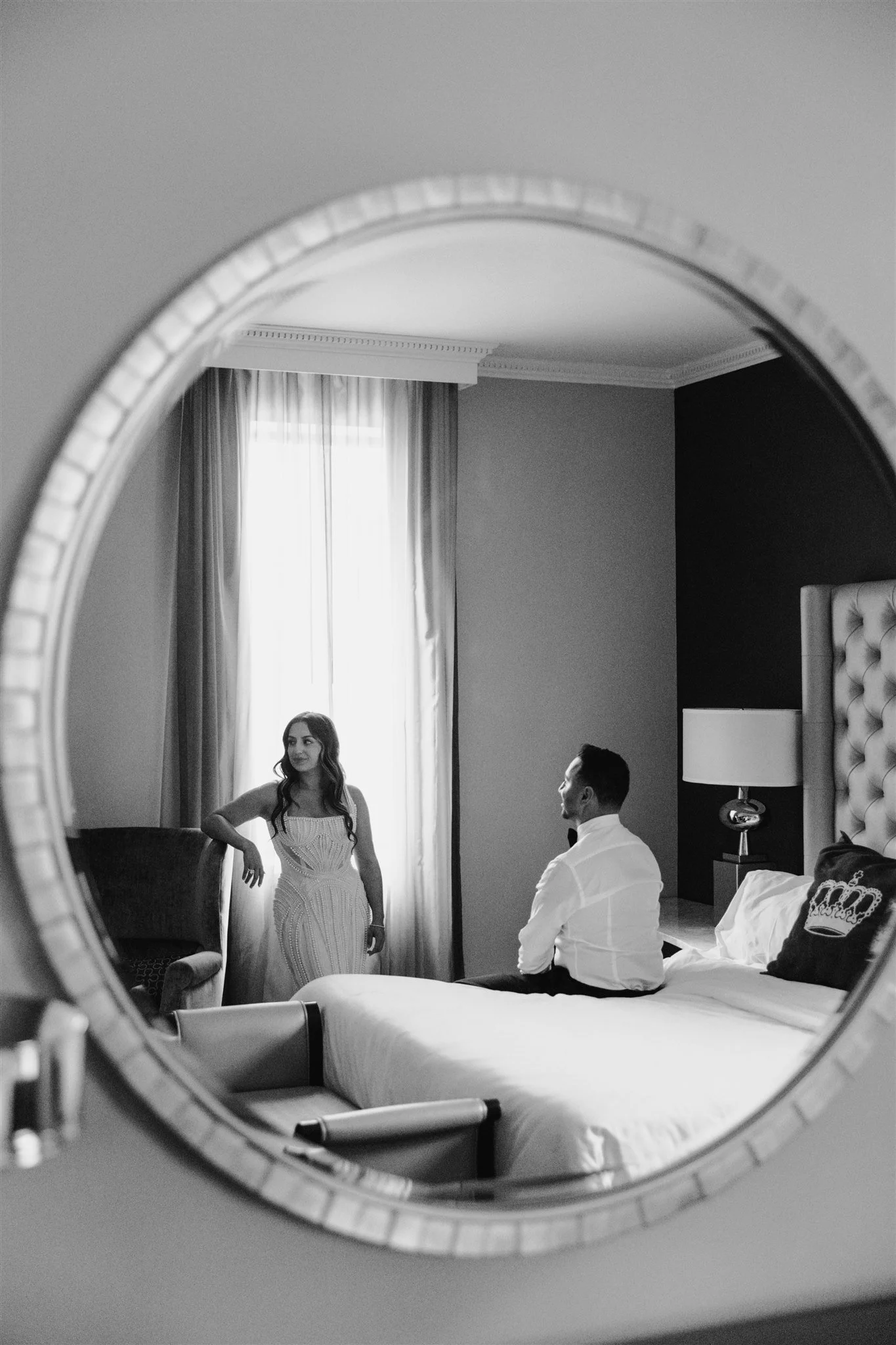 A black and white photo seen through a round mirror showing a woman in a dress standing in a room with a man sitting on a bed, with a window with curtains in the background.
