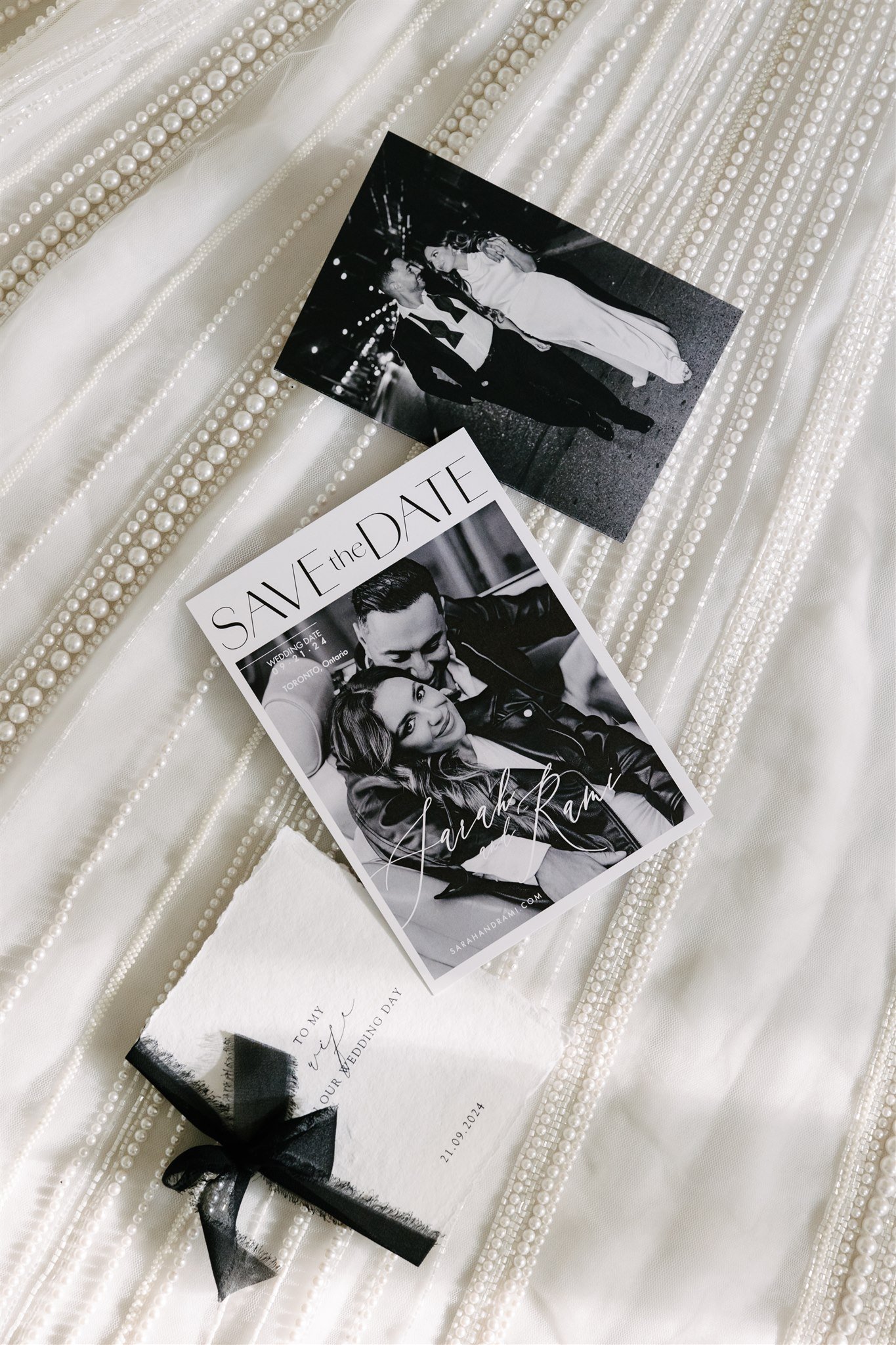 Wedding save the date card with a photo of a couple and a smaller card with a wedding date and a bow on it, placed on a pearl-embellished fabric background.