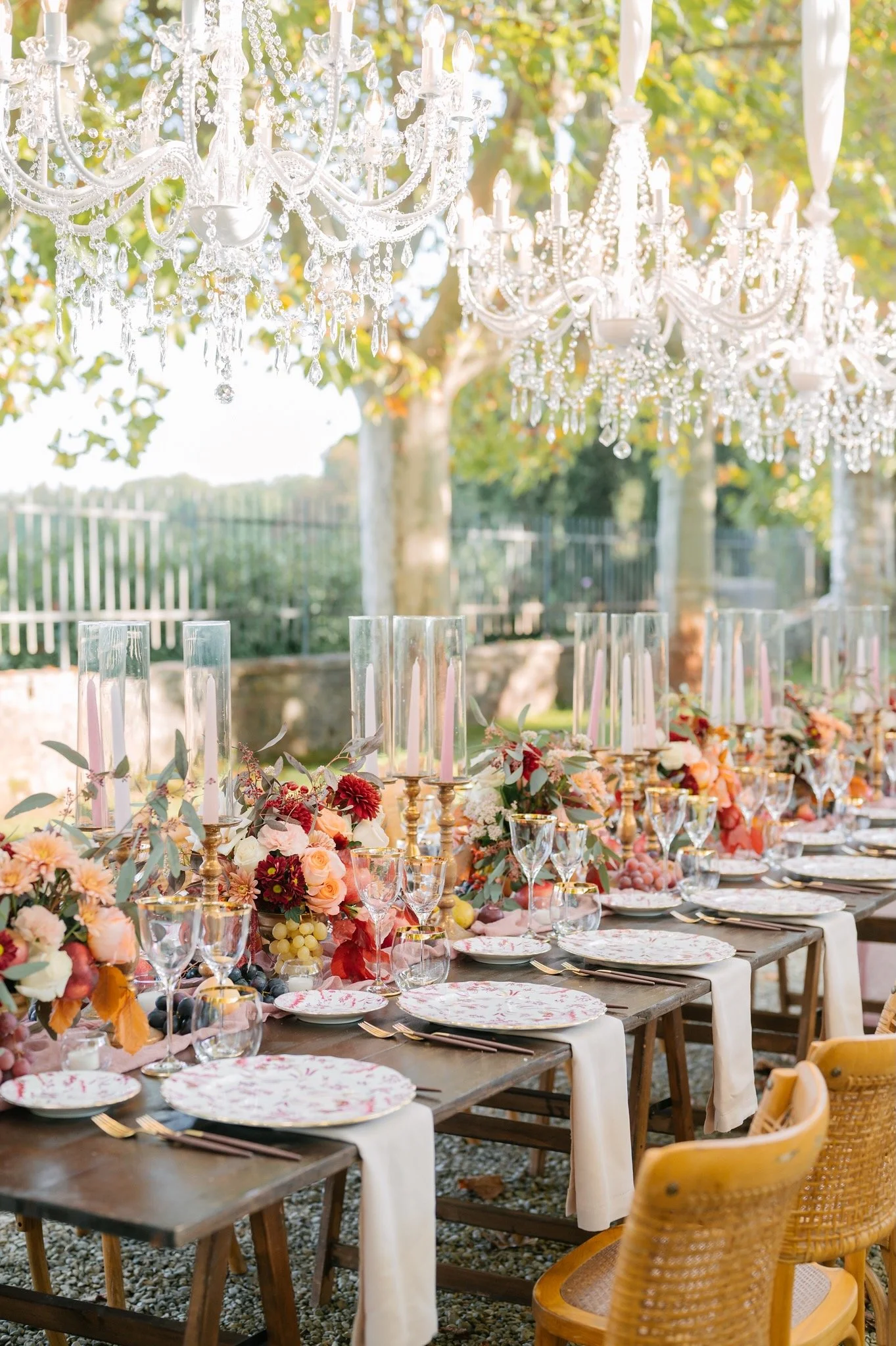 Outdoor wedding table setup with floral centerpieces, candles, and elegant tableware, under crystal chandeliers.