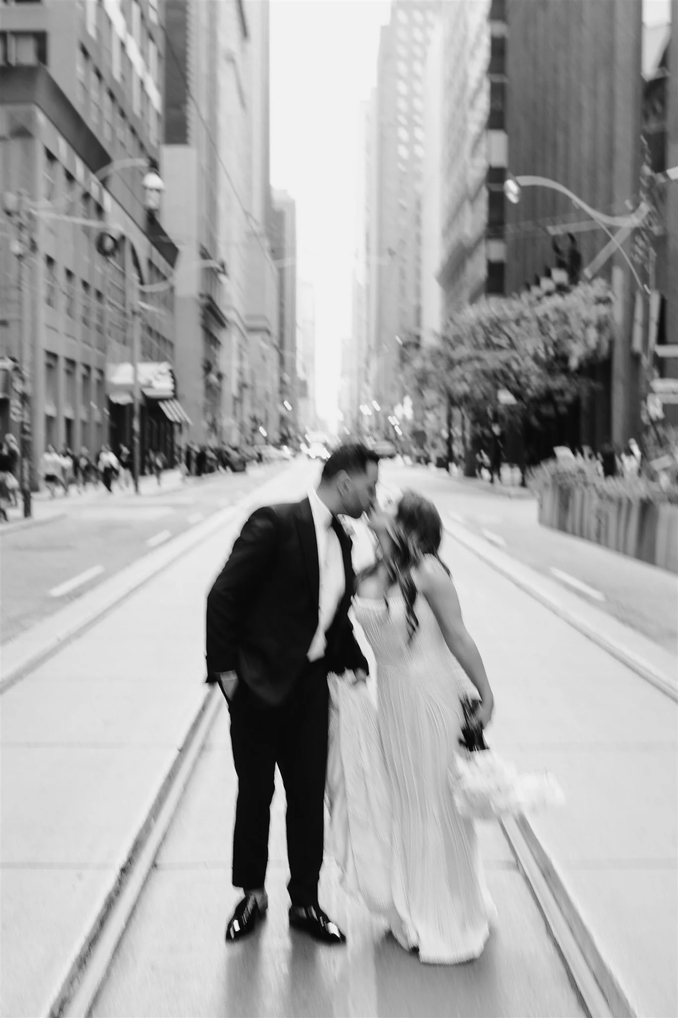 A black and white photo capturing a couple kissing in the middle of a city street surrounded by tall buildings, with some people visible in the background.