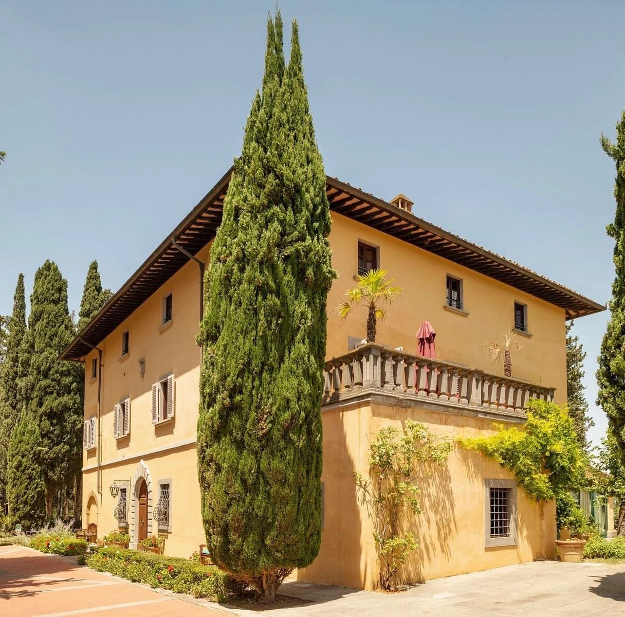 A two-story yellow Mediterranean-style house with a tiled roof, surrounded by tall green cypress trees, with a balcony and potted plants, set against a clear blue sky.
