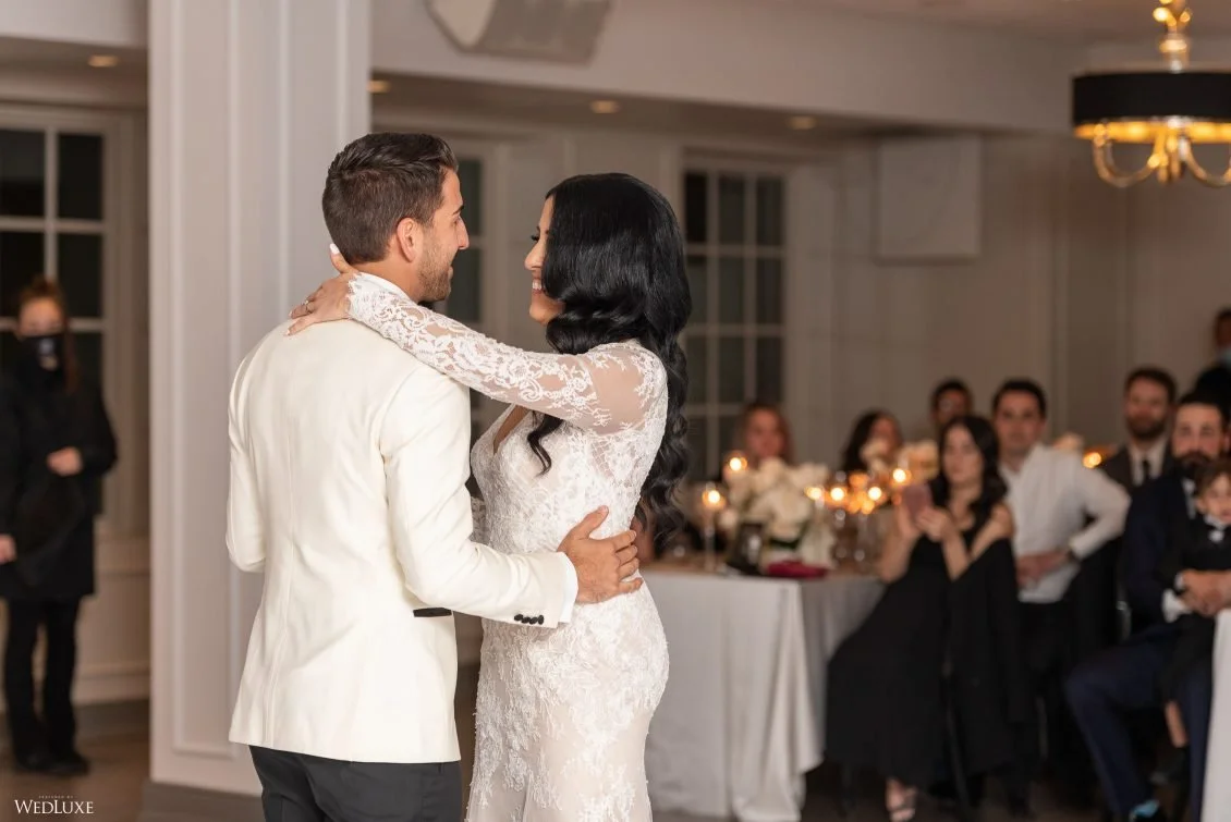 A bride and groom dance together at their wedding reception, surrounded by seated guests in a warmly lit room.