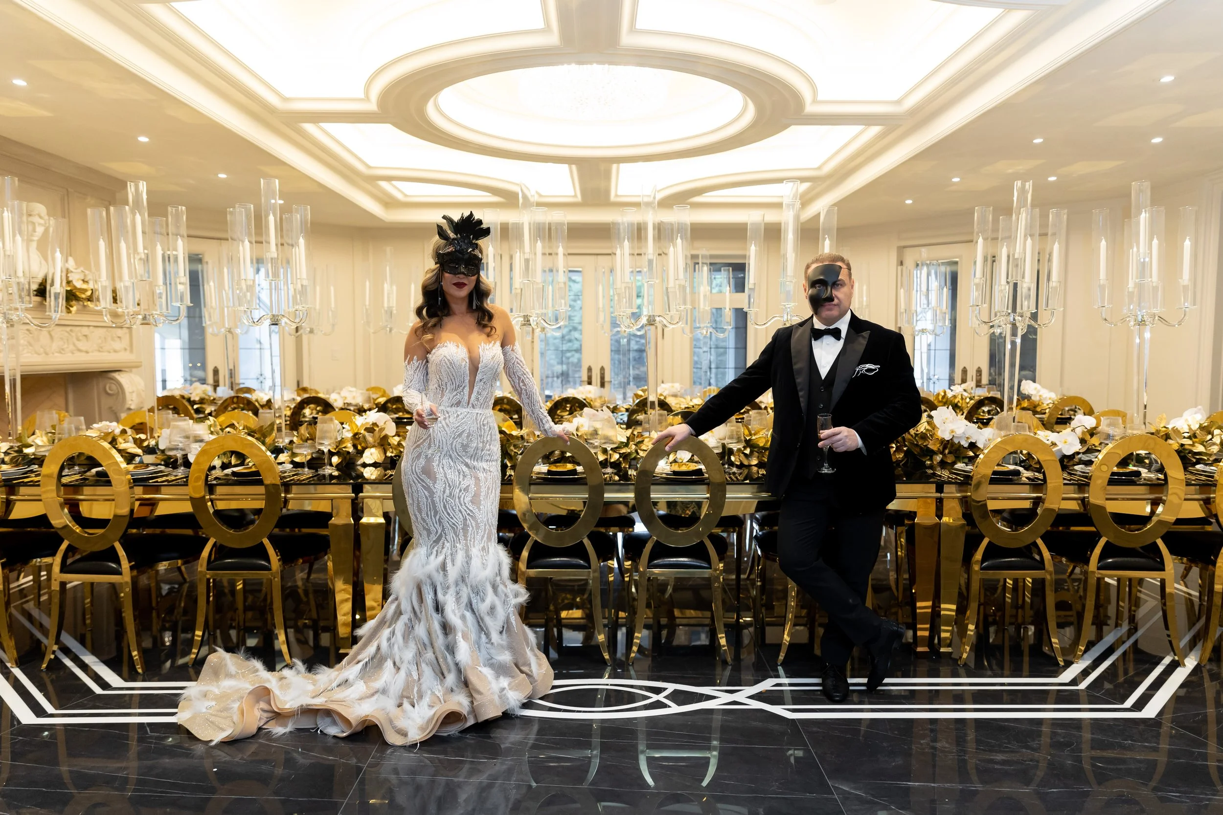 A man and woman dressed in formal attire, wearing masks, standing in front of an elegantly decorated dining table with gold accents and tall candelabras in a luxurious, well-lit room.