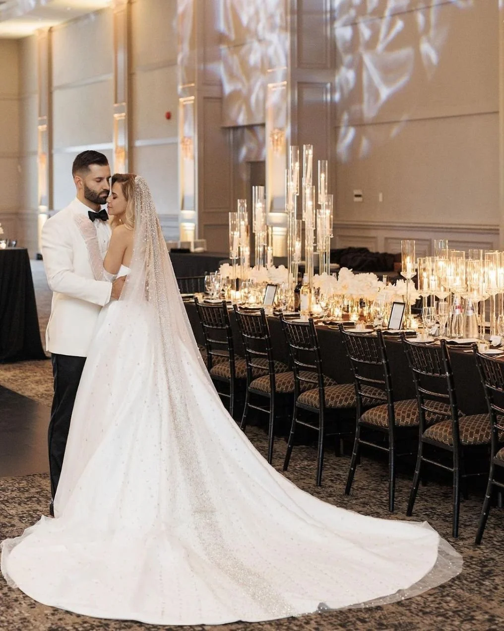 A bride and groom embrace in a wedding reception hall with elegant decor and a long decorated table.
