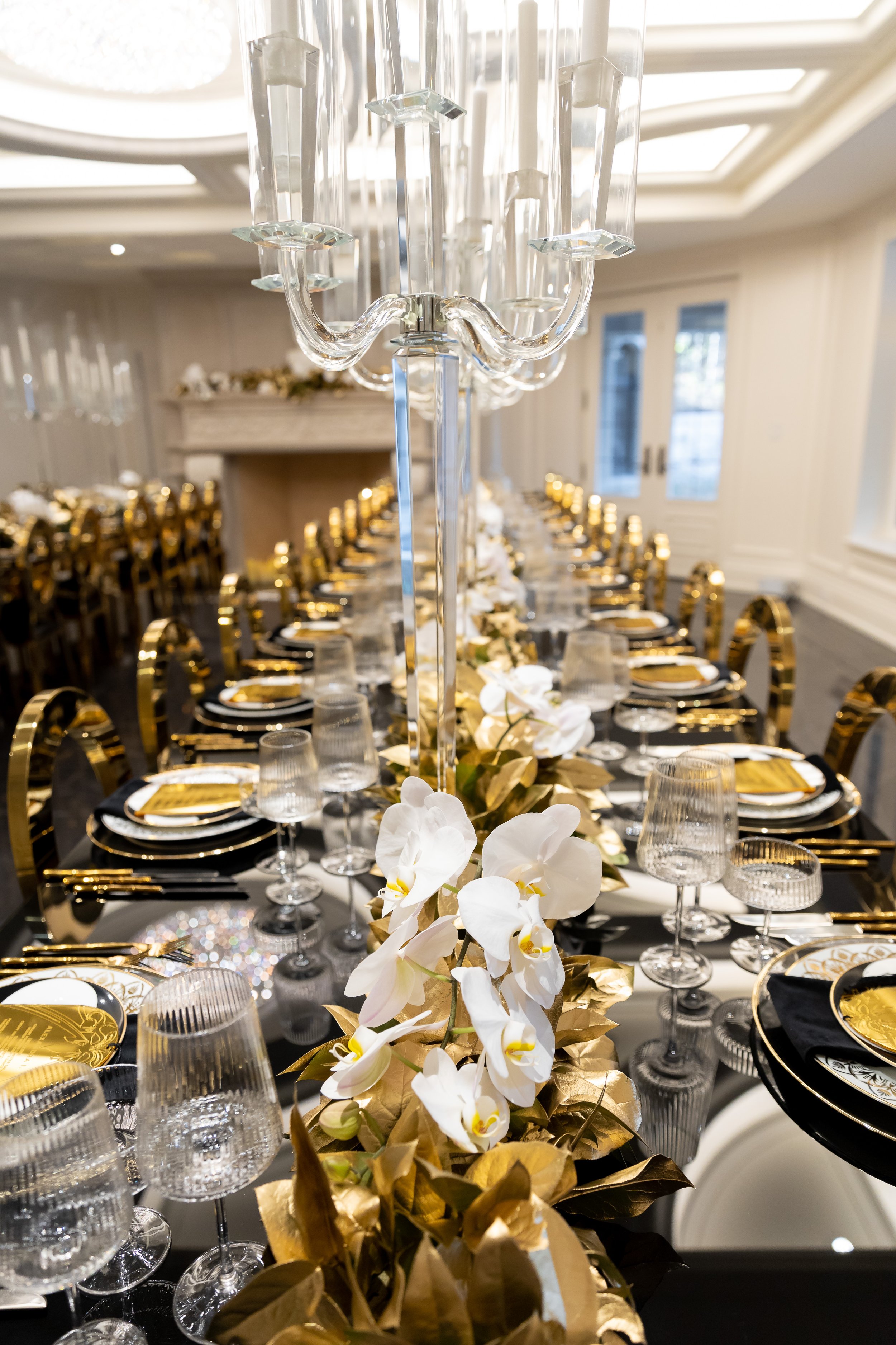 A long banquet table decorated with white orchids and gold accents, set with black and gold plates, multiple wine glasses, silverware, and a tall crystal candelabrum in a luxurious room.