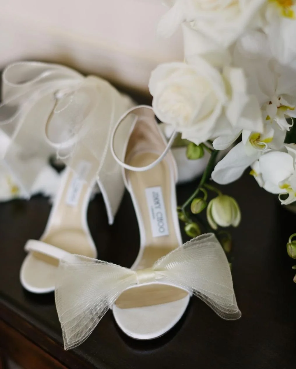 Cream-colored high heel shoes with a large bow on the front, placed next to a bouquet of white flowers.