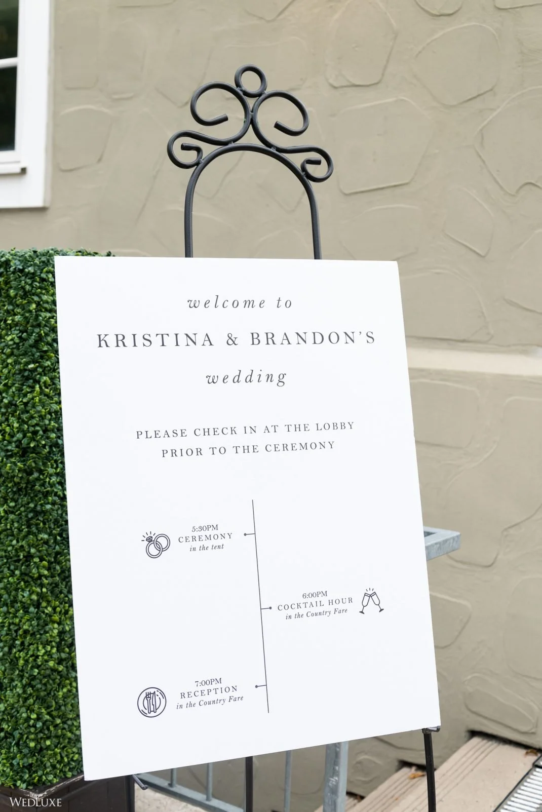 Wedding schedule sign for Kristina and Brandon's wedding with ceremony at 5:30 PM, cocktail hour at 6:00 PM, and reception at 7:00 PM, displayed on an easel outside with greenery and a stone wall in the background.
