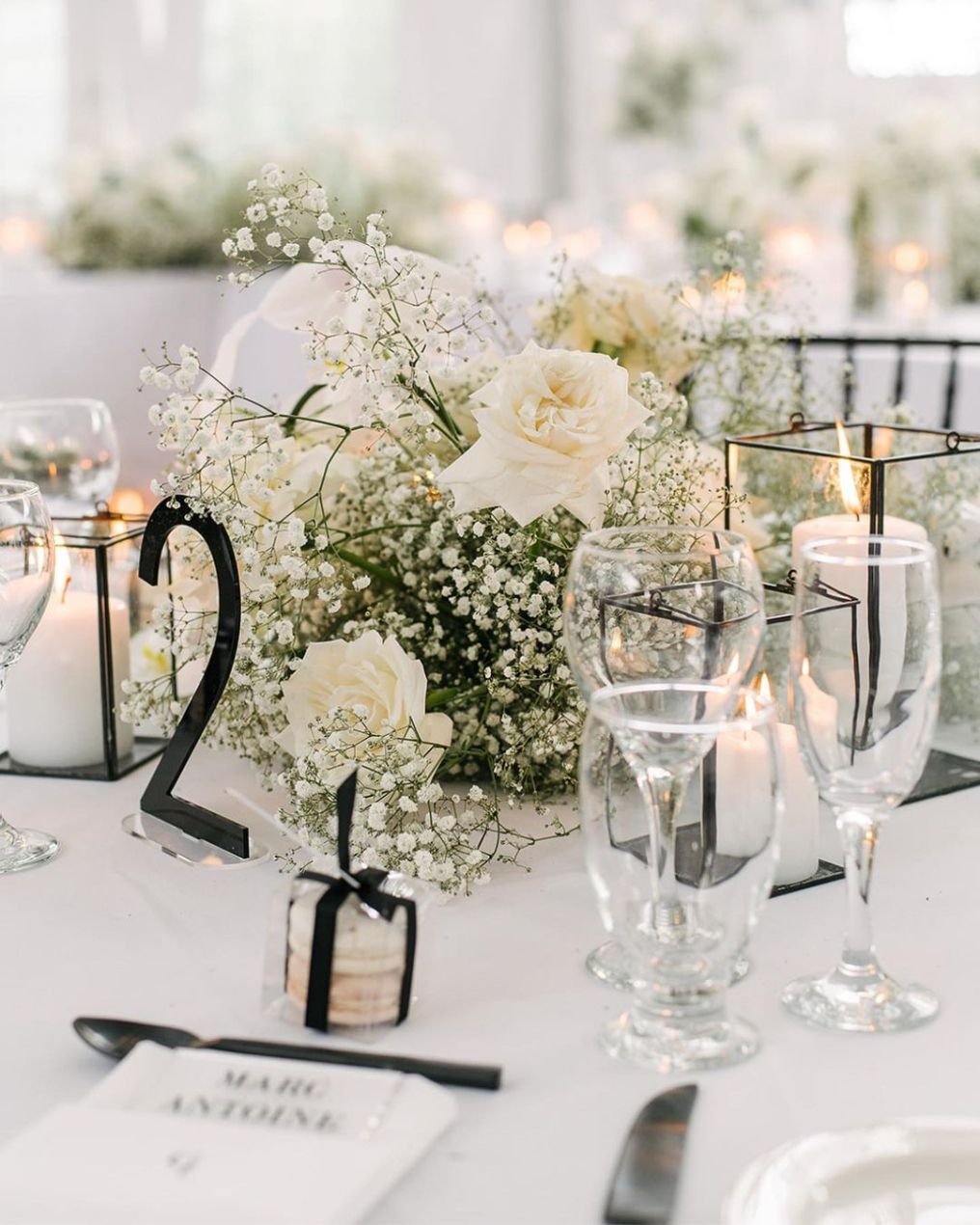 Elegant wedding table centerpiece with white roses, baby's breath, candles in glass containers, and table numbers.