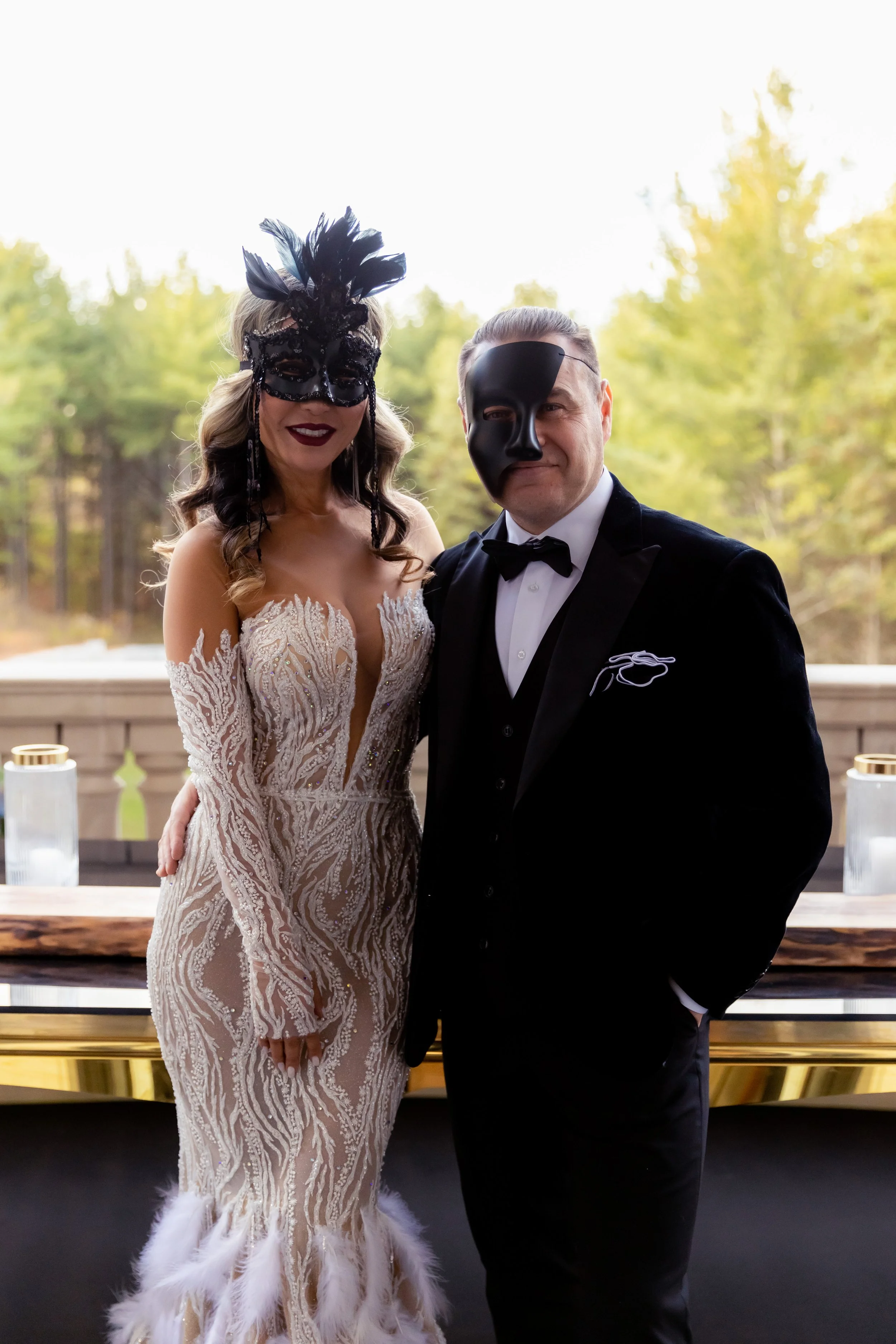A woman in an ornate wedding dress and a woman in a black tuxedo wearing masquerade masks at a formal event.