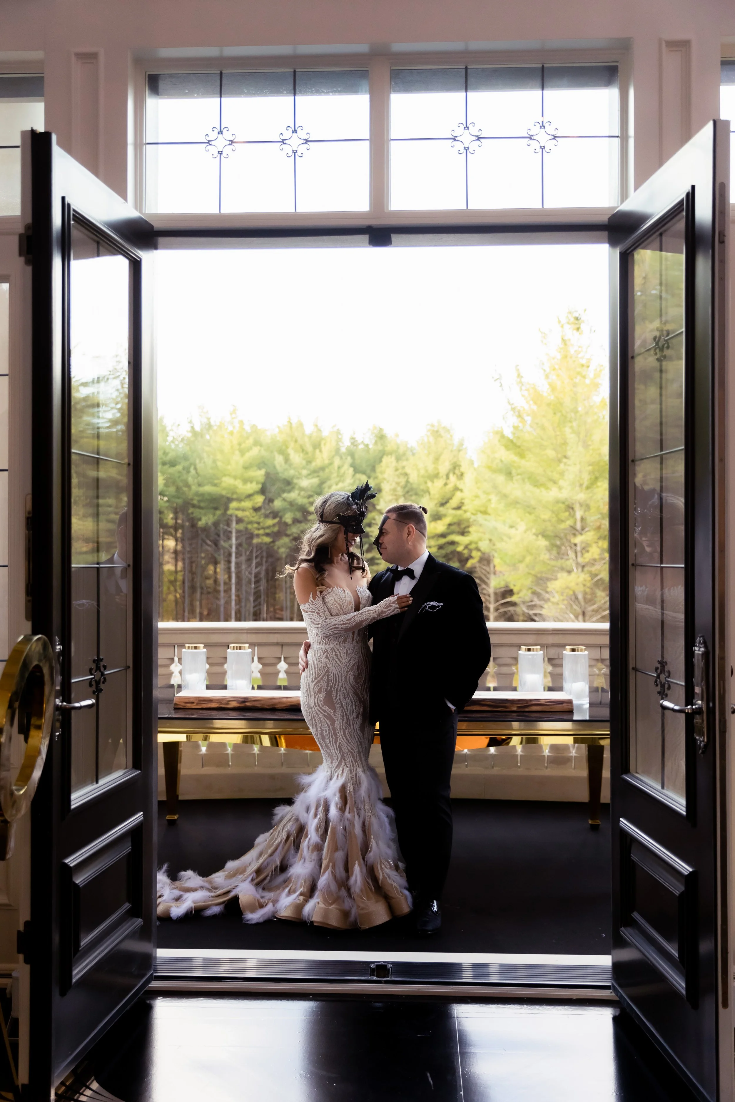 A bride and groom standing on a balcony, framed by open double doors, with trees in the background.