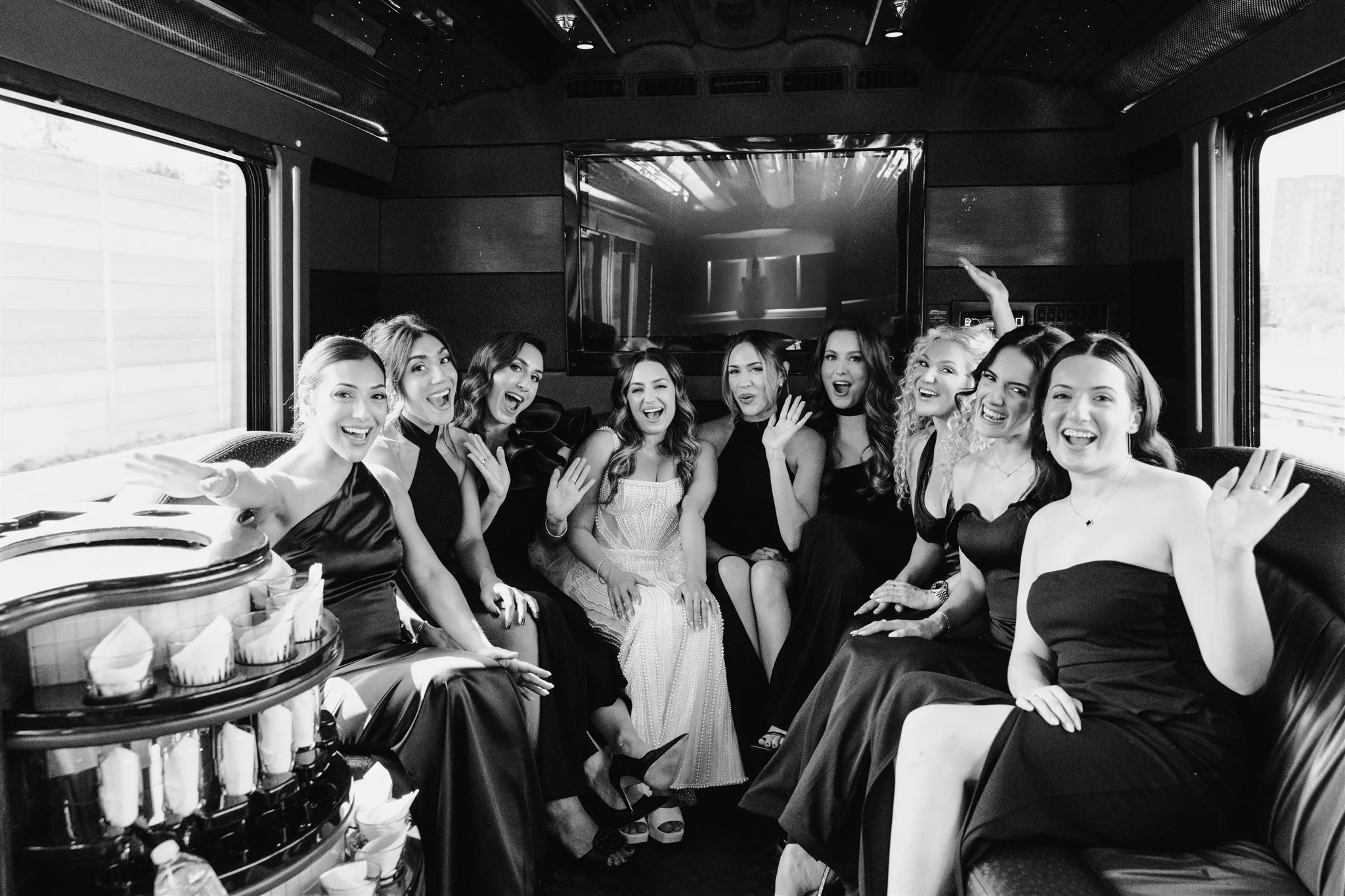 A group of women dressed in evening gowns sitting inside a luxurious vehicle, smiling and waving at the camera.