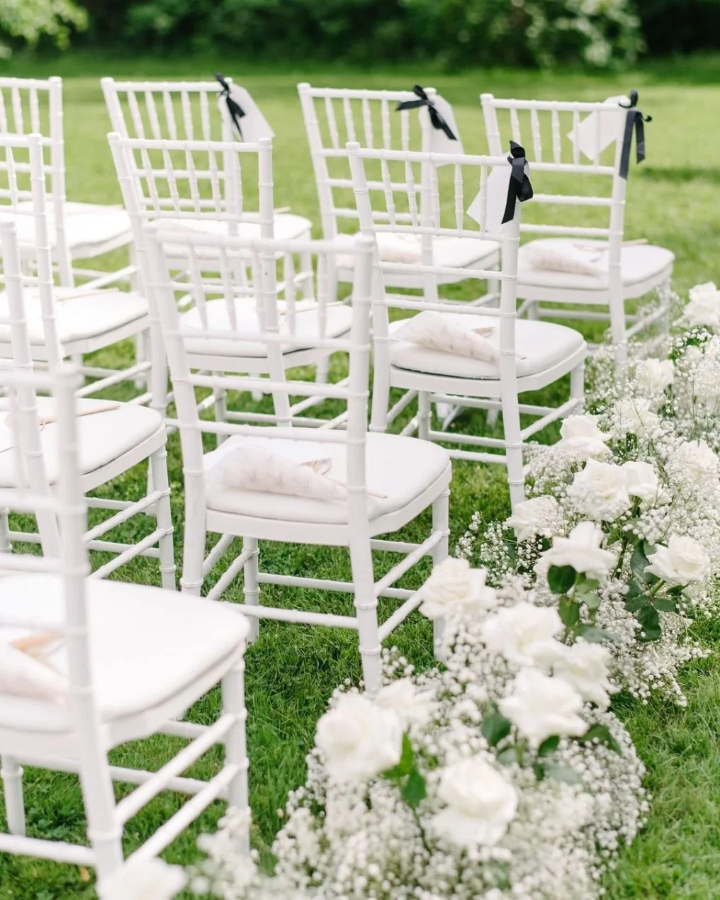 White chairs arranged in rows on a grassy outdoor setting, decorated with black ribbons, with white flowers in the foreground, suggesting a wedding setup.