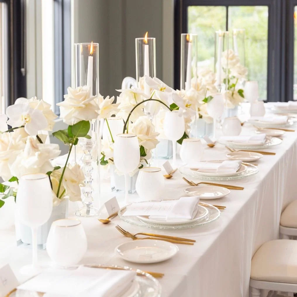 Elegant wedding or event table with white floral centerpieces, candles, white tableware, and gold utensils.