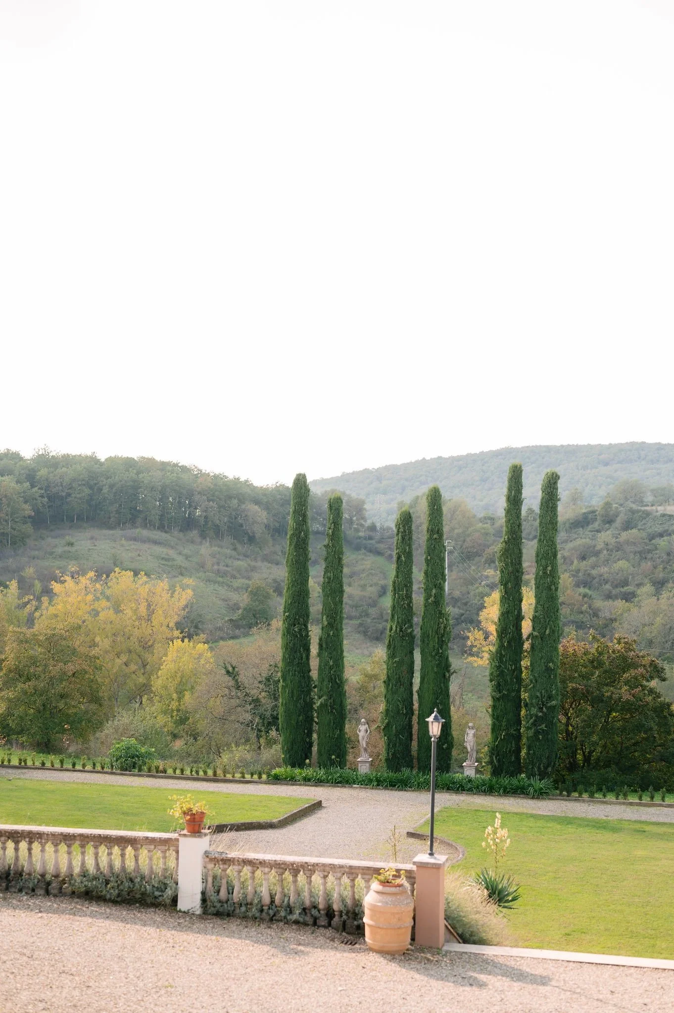 Scenic view of a landscaped garden with tall cypress trees, statues, a lamp post, and potted plants, set against a backdrop of rolling hills and lush greenery.