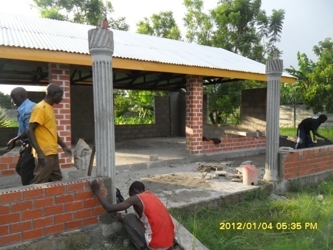 This is the former front view of Dr. Elizabeth Rini Medical Center before roofing the front Corridor. From (L –R) was Stanley Mpuku (in blue), Uncle Sam (in yellow,) Cliff (in red) and on the far corner of the corridor was Isaac building a shield of 70 centimeters high around the front corridor from one pillar to the other using donated pan bricks (red bricks) leaving space for the entrance