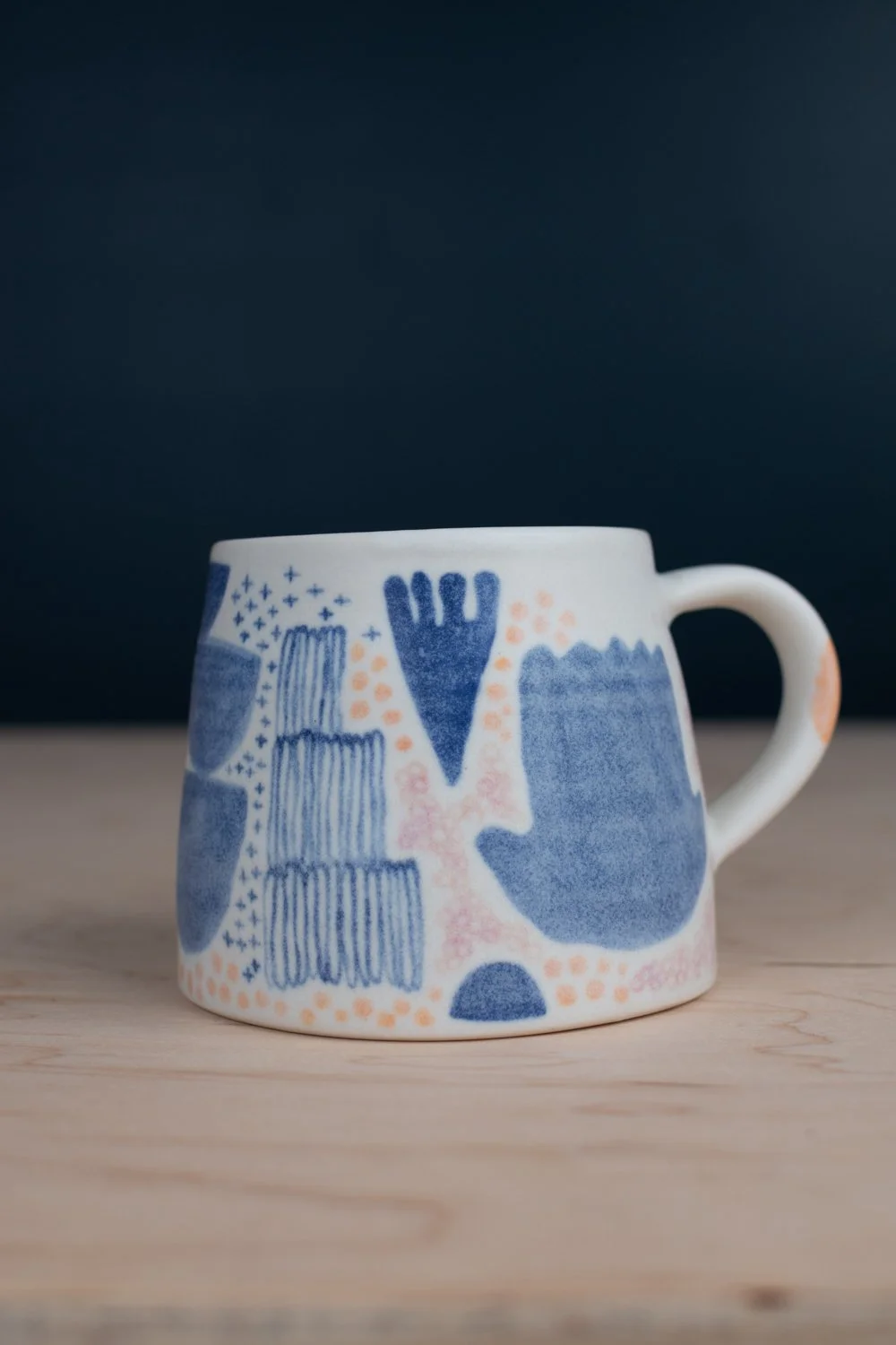 Hand-thrown porcelain mug with blue, blush, and tangerine illustrations painted on the surface.