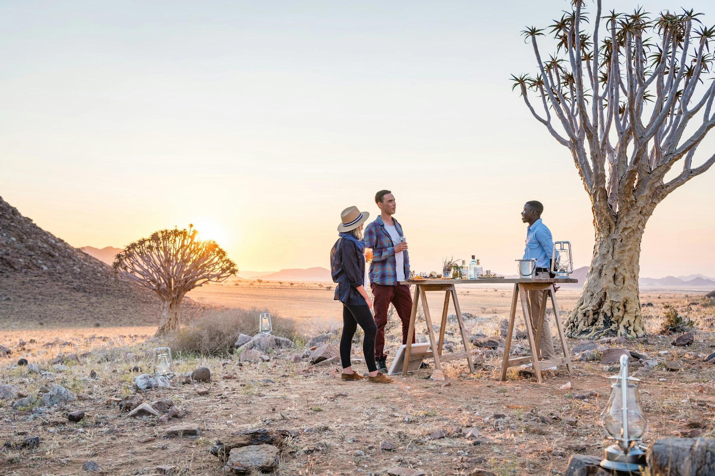 Three people standing around a portable outdoor bar in a desert landscape at sunset, with a large desert tree and smaller trees in the background.