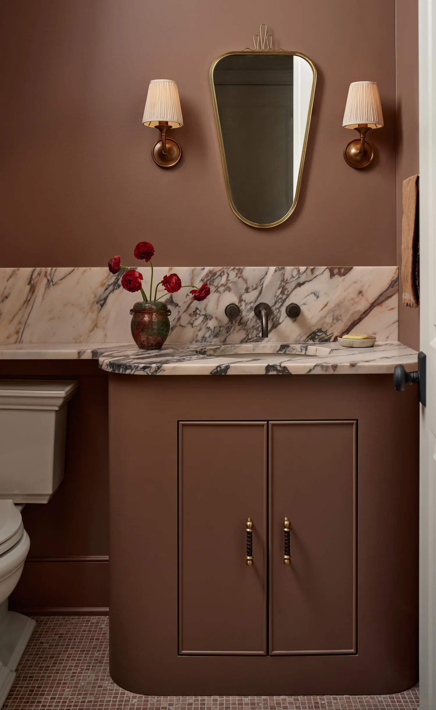 A bathroom with a brown wall, a marble countertop, a mirror, two wall sconces, a vase with red flowers, and a cabinet with gold and black handles.