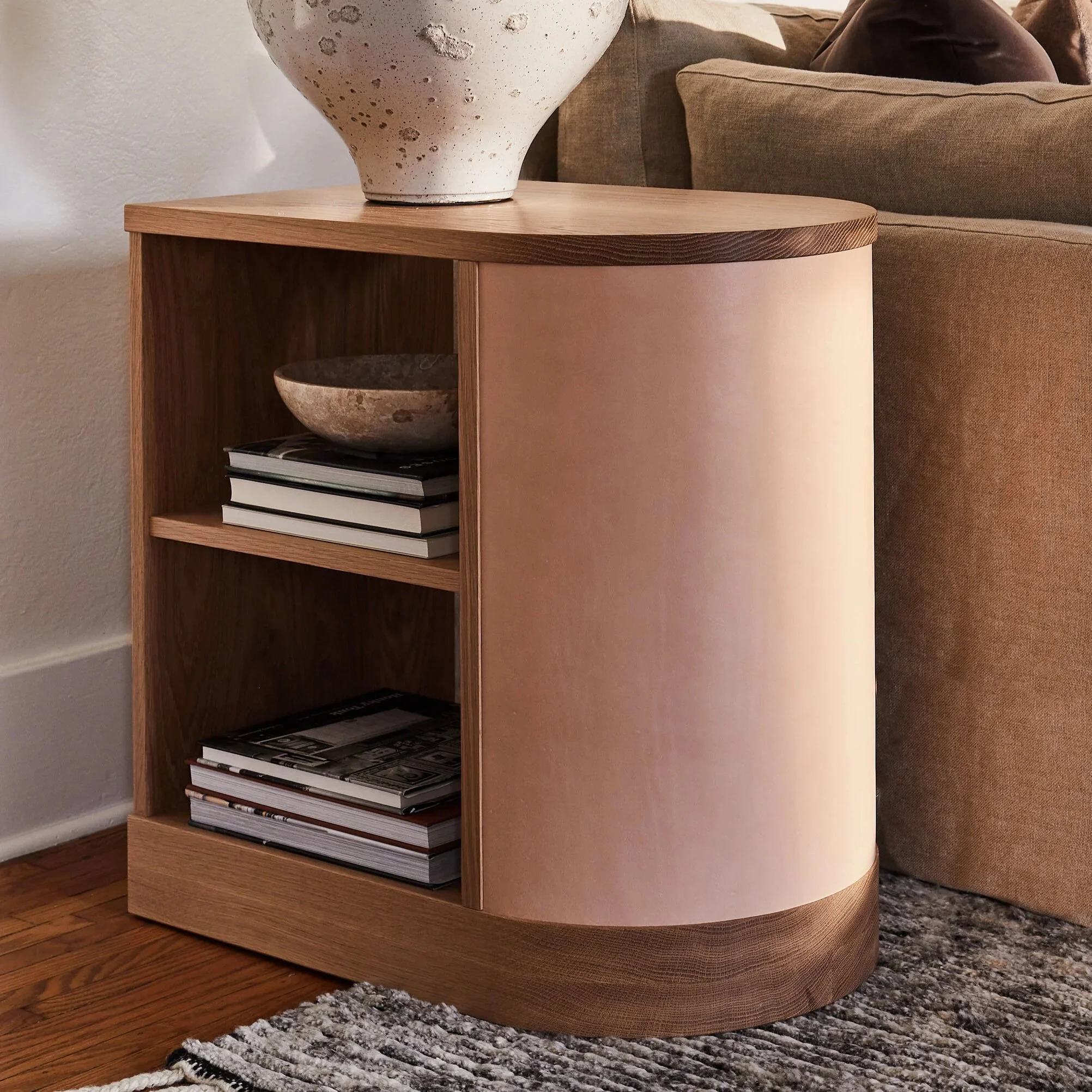 A modern wooden side table with built-in shelves, positioned next to a beige fabric sofa on a textured gray and black rug, with a decorative large white ceramic vase on top of the table in a cozy living room.