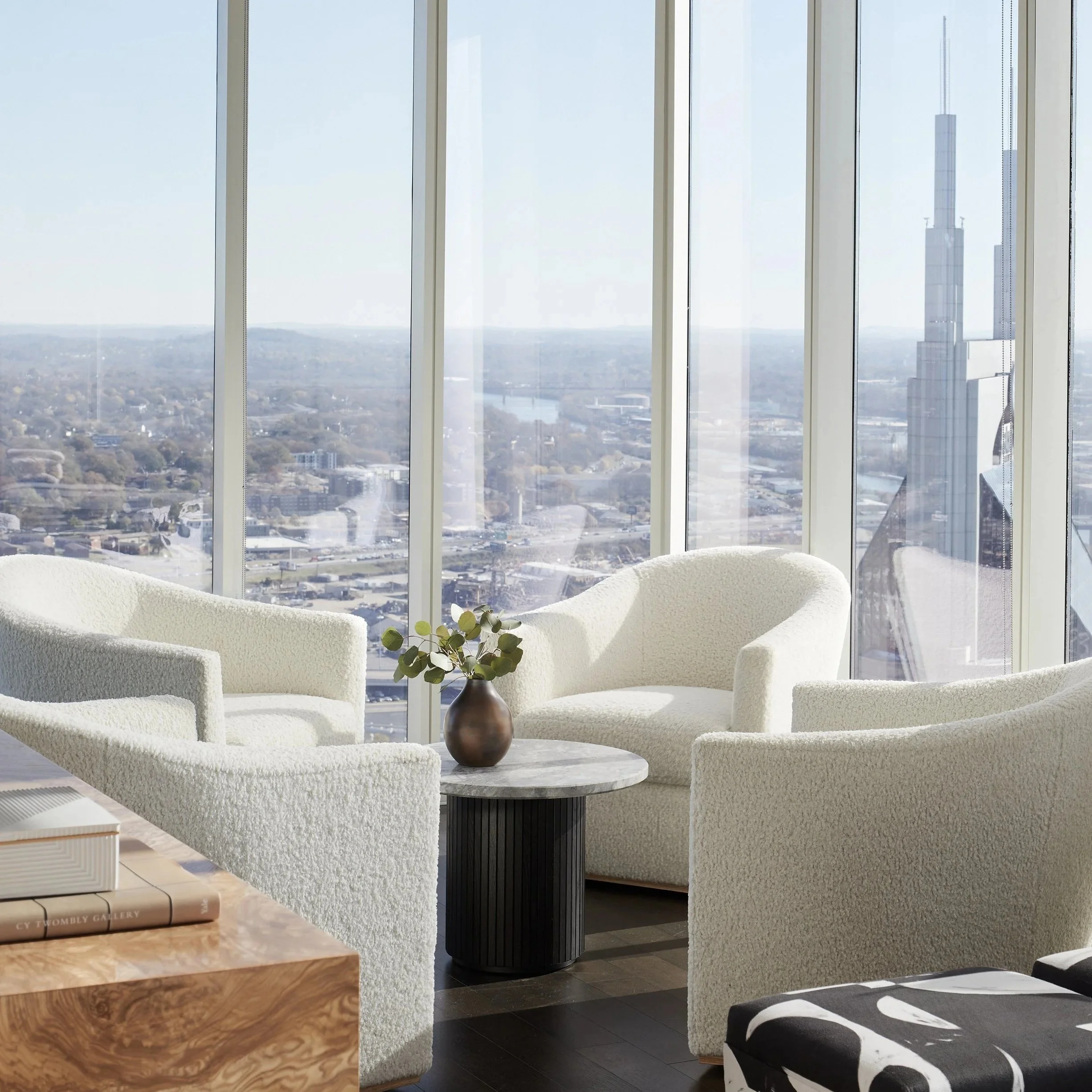 Modern living room with plush white sofas, a black cylindrical side table, and large floor-to-ceiling windows showcasing a city skyline with tall buildings and a river in the background.