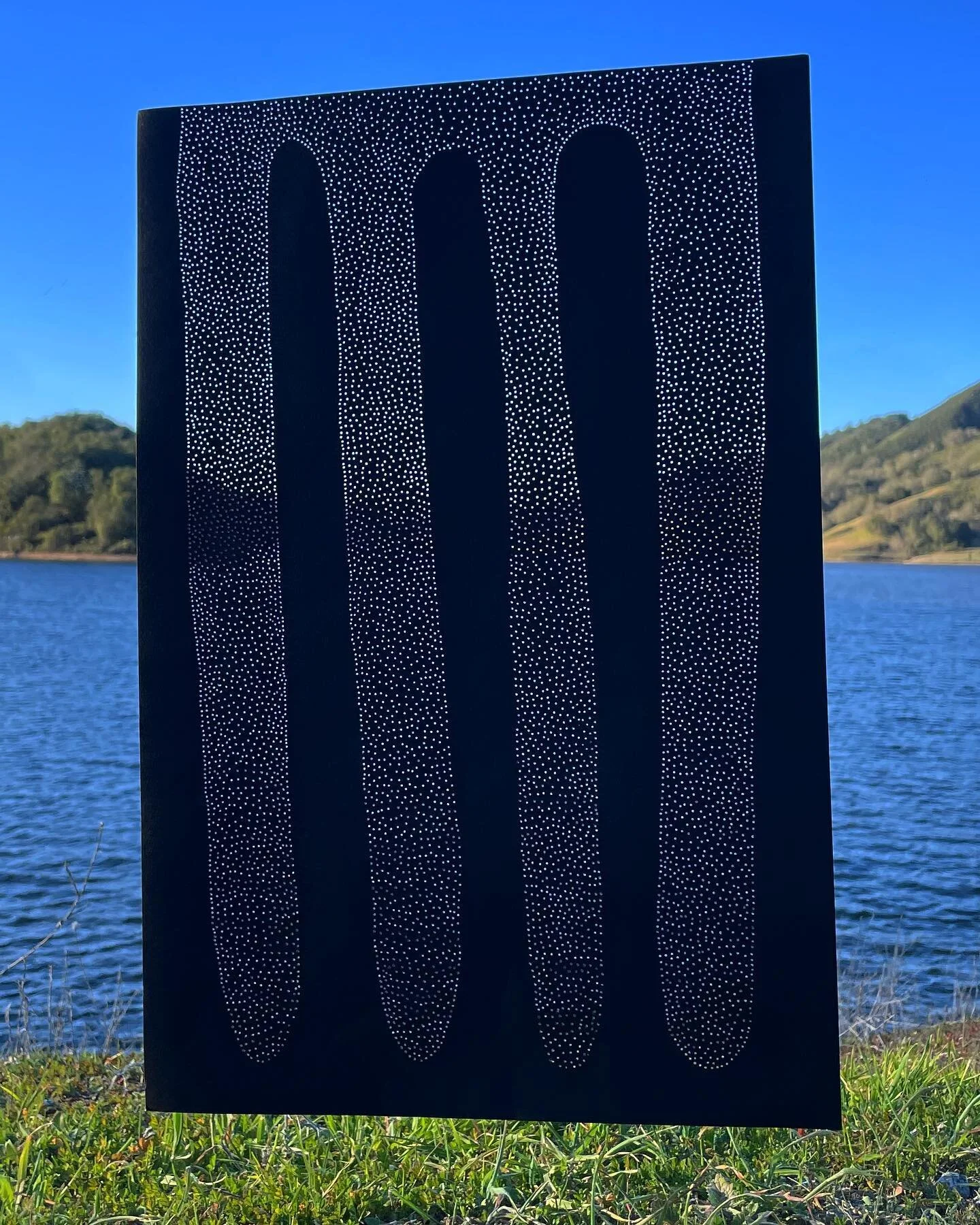 works on paper β β‘ drawings for a window 
ID: a sheet of black paper floats in front of a blue sky, rolling hills, navy blue water, and green grass. a black squiggle, shaped like three long fingers, moves its way up the paper interlocking with anothe