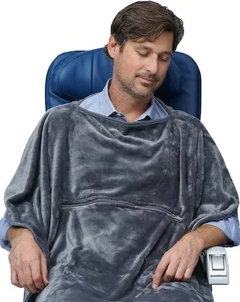 Man resting on a plane seat wearing a poncho-style travel blanket with front pocket