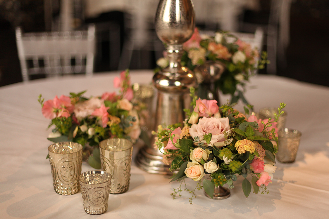 Garden centerpieces in the Continental Room at the Hilton Netherland Plaza Hotel, by Cincinnati wedding florist Floral Verde LLC. Centerpiece contains coral sweet peas, apple blossom yarrow, Ambridge garden roses, Chablis spray roses, apricot stock, Quicksand roses, White Majolica spray roses, white larkspur, mint, scented geranium, lemon basil, bupleurum, seeded eucalyptus and Artemisia 'Powis Castle' in vintage silver, surrounded by mercury glass votives.