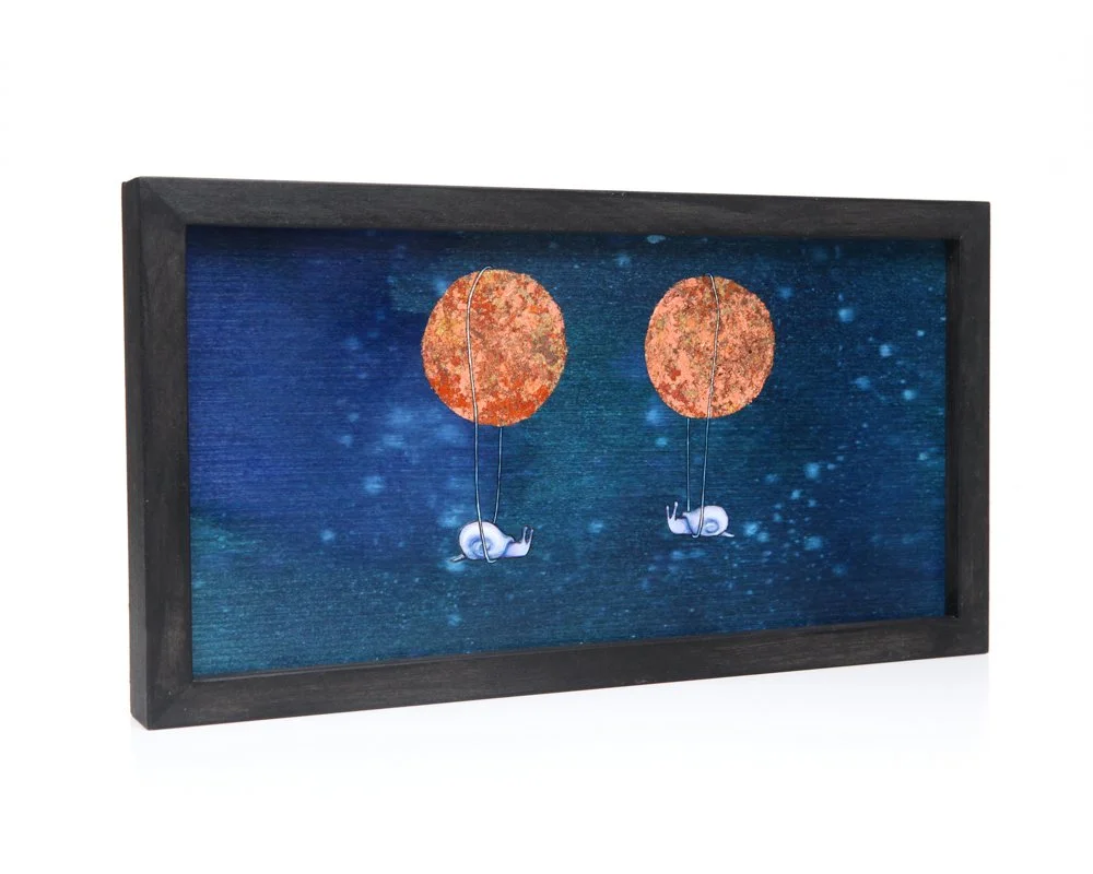 A framed artwork features two snails holding strings attached to moons with hints of gold leaf, set against a deep blue, starry night sky