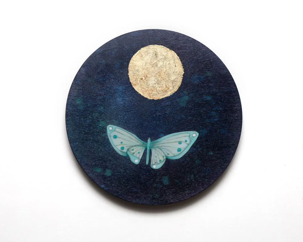 A circular artwork features a golden moon above a light blue moth with spotted wings, against a deep blue, star-speckled night sky. The scene is simple with the moth flying toward the glowing moon.