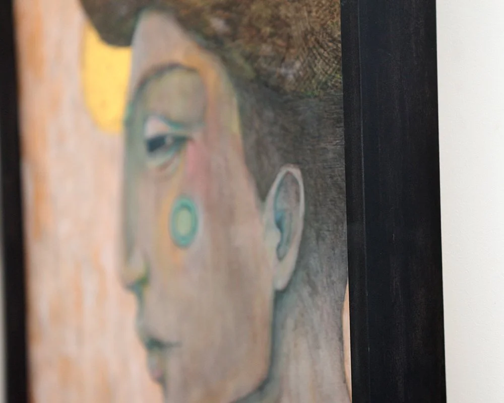 A side profile of a person with greenish skin tones, prominent cheek and chin, and high textured brown hair. They have a blue circle on their cheek and a gold crescent shape above the eye