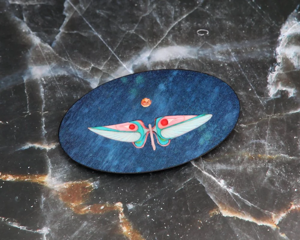 An oval artwork with a blue textured background by Kate Morgan Art features a stylized moth in pastel blue, pink, and teal at the center, with a small golden moon above its head.