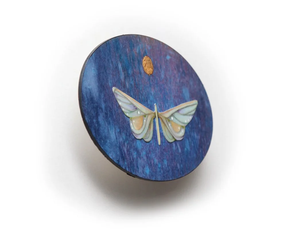 An oval-shaped artwork by Kate Morgan Art features a stylized white moth with outspread wings against a textured blue-purple background. Above the moth, a small gold circle resembles the moon.