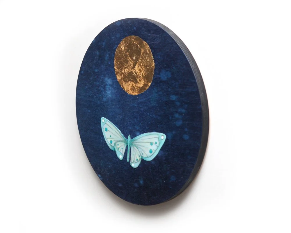 A circular artwork features a golden moon above a light blue moth with spotted wings, against a deep blue, star-speckled night sky. The scene is simple with the moth flying toward the glowing moon.