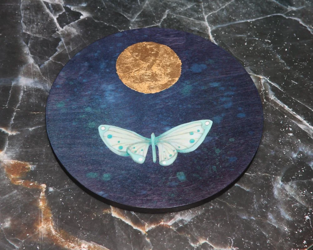 A circular artwork features a golden moon above a light blue moth with spotted wings, against a deep blue, star-speckled night sky. The scene is simple with the moth flying toward the glowing moon.