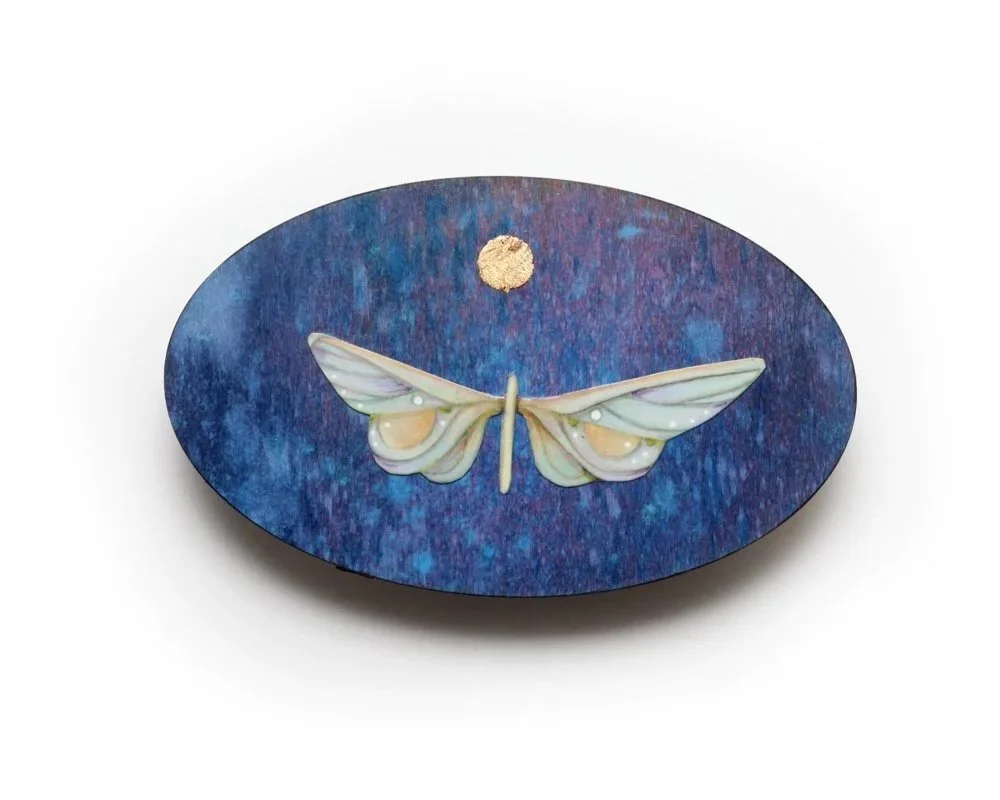 An oval-shaped artwork by Kate Morgan Art features a stylized white moth with outspread wings against a textured blue-purple background. Above the moth, a small gold circle resembles the moon.