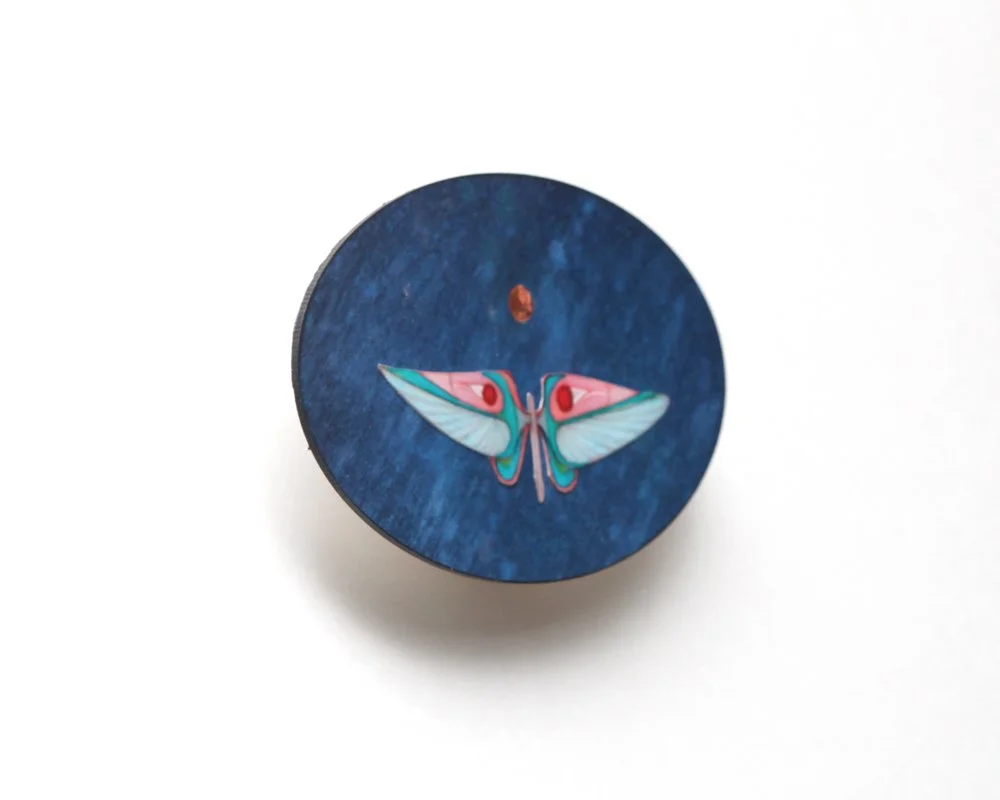 An oval artwork with a blue textured background by Kate Morgan Art features a stylized moth in pastel blue, pink, and teal at the center, with a small golden moon above its head