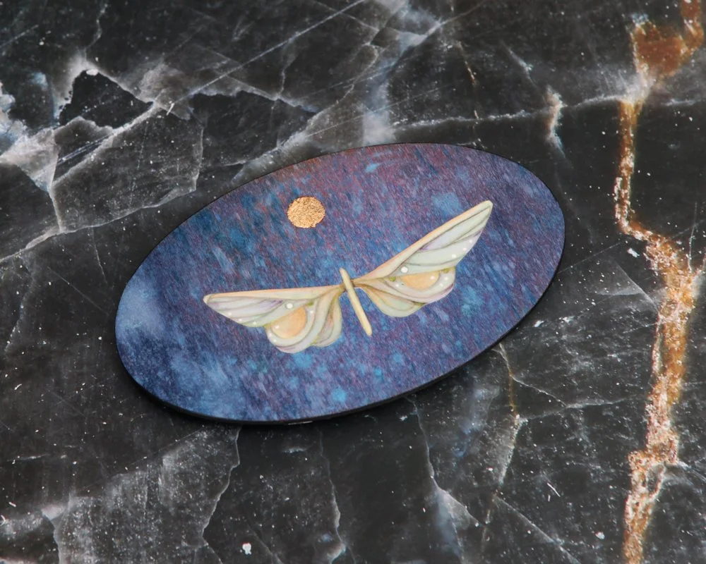 An oval-shaped artwork by Kate Morgan Art features a stylized white moth with outspread wings against a textured blue-purple background. Above the moth, a small gold circle resembles the moon.