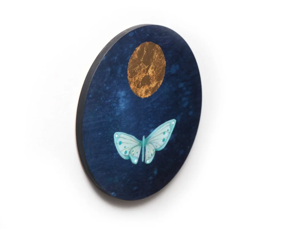 A circular artwork features a golden moon above a light blue moth with spotted wings, against a deep blue, star-speckled night sky. The scene is simple with the moth flying toward the glowing moon.