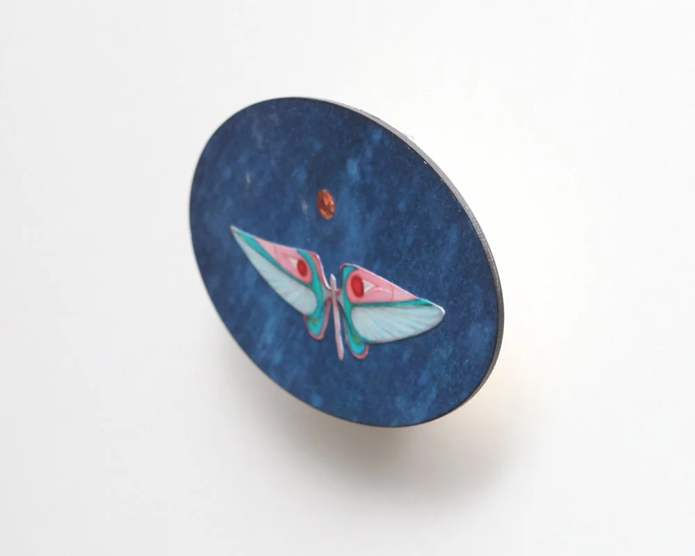 An oval artwork with a blue textured background by Kate Morgan Art features a stylized moth in pastel blue, pink, and teal at the center, with a small golden moon above its head
