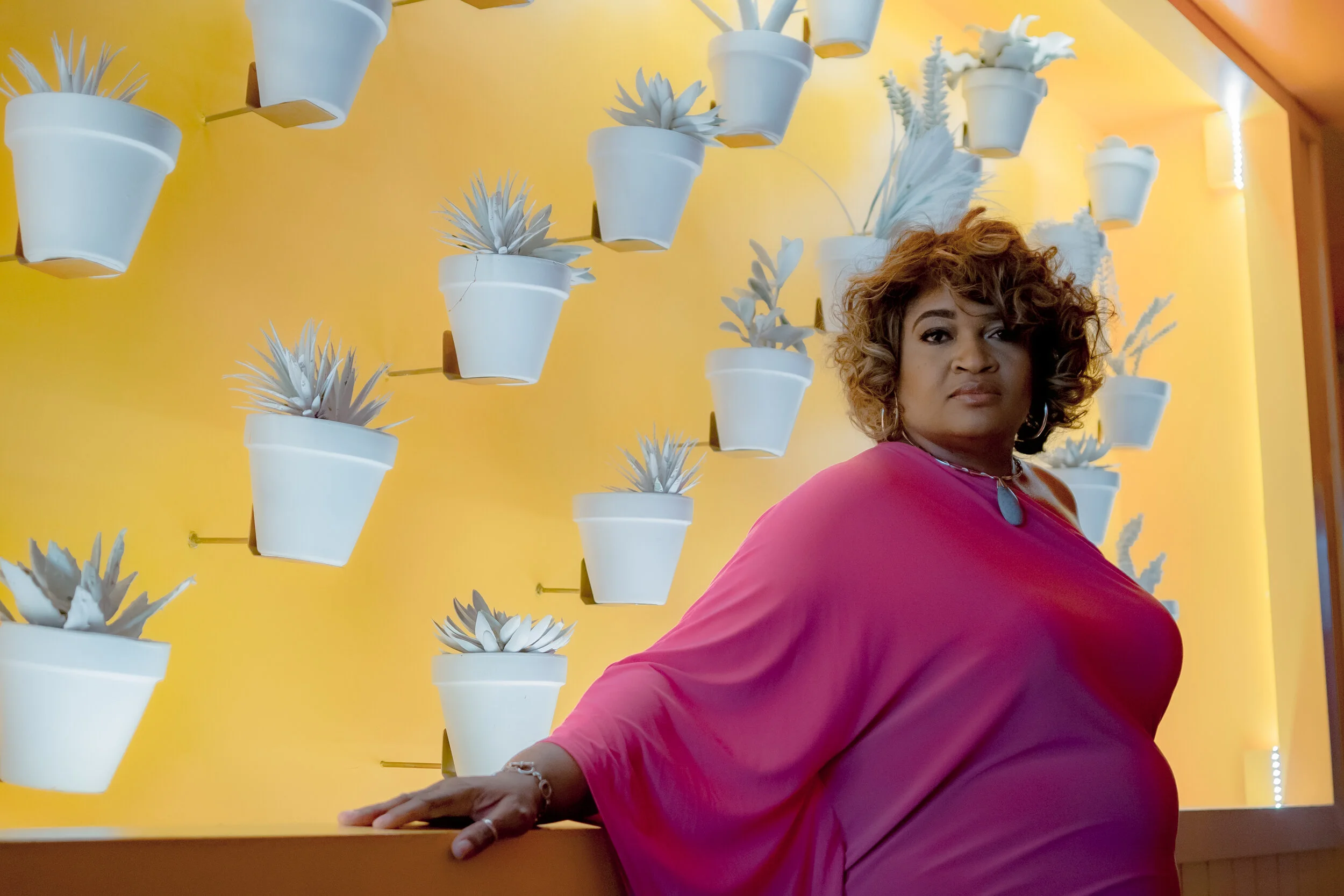 A woman in a pink dress standing in front of a yellow wall decorated with white potted succulents.