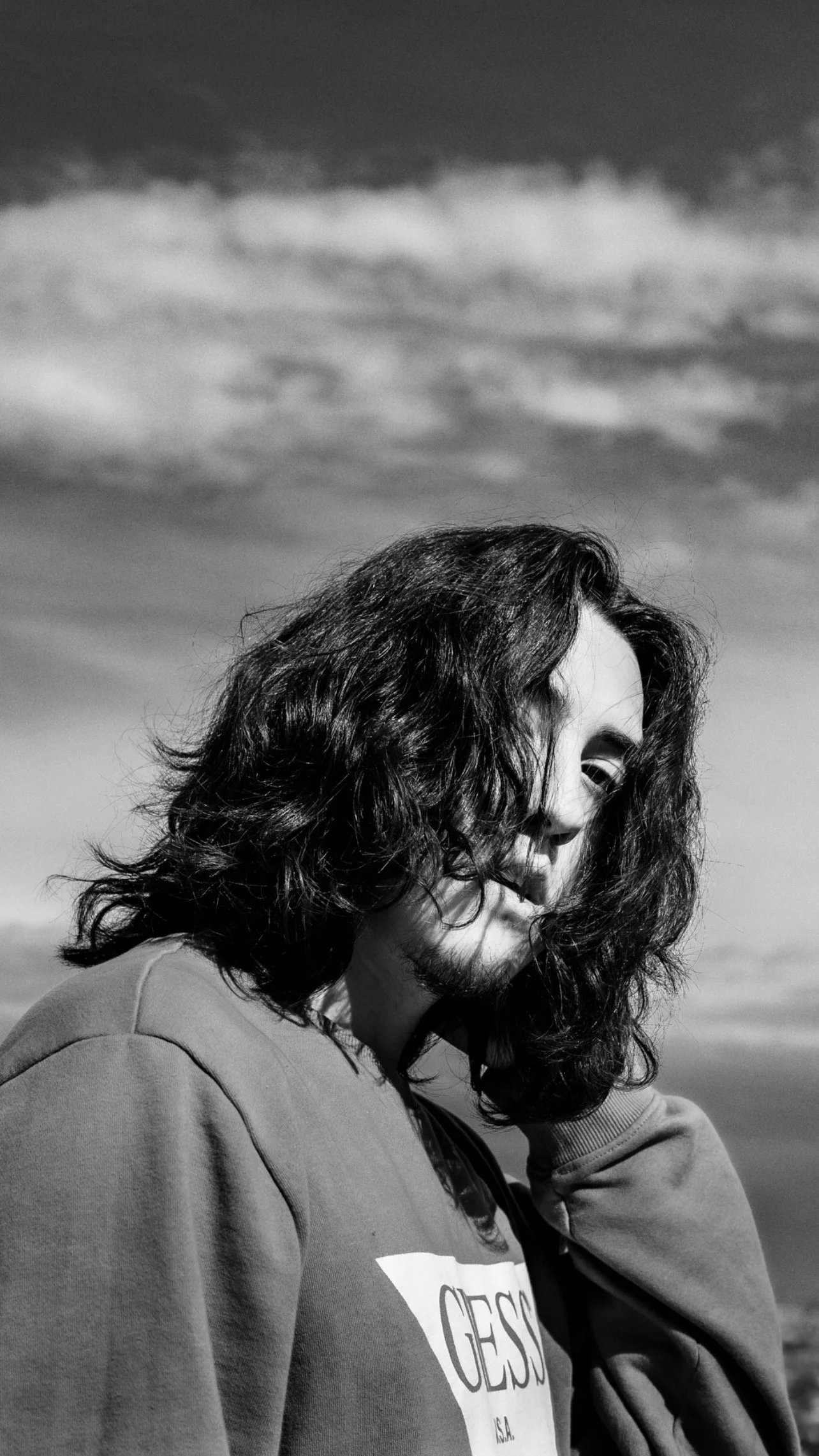 Black-and-white photo of a person with shoulder-length curly hair smiling, wearing a hoodie, outdoors with cloudy sky background.