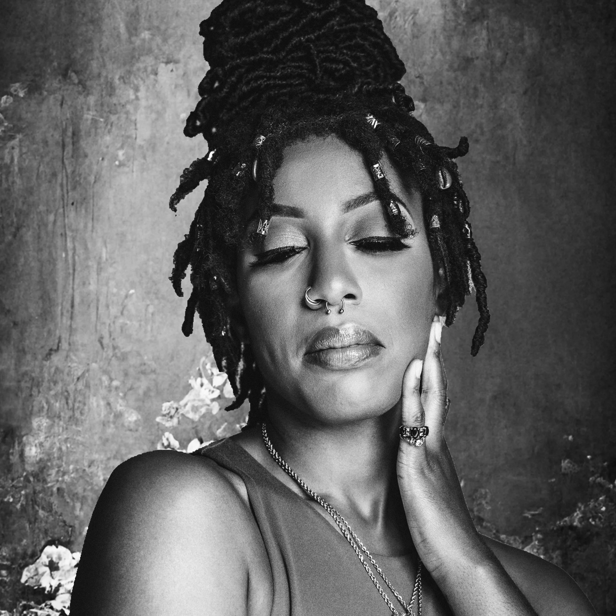 A black and white portrait of a woman with dreadlocks styled in an updo, closed eyes, and hand resting on her face, wearing jewelry and a sleeveless top.