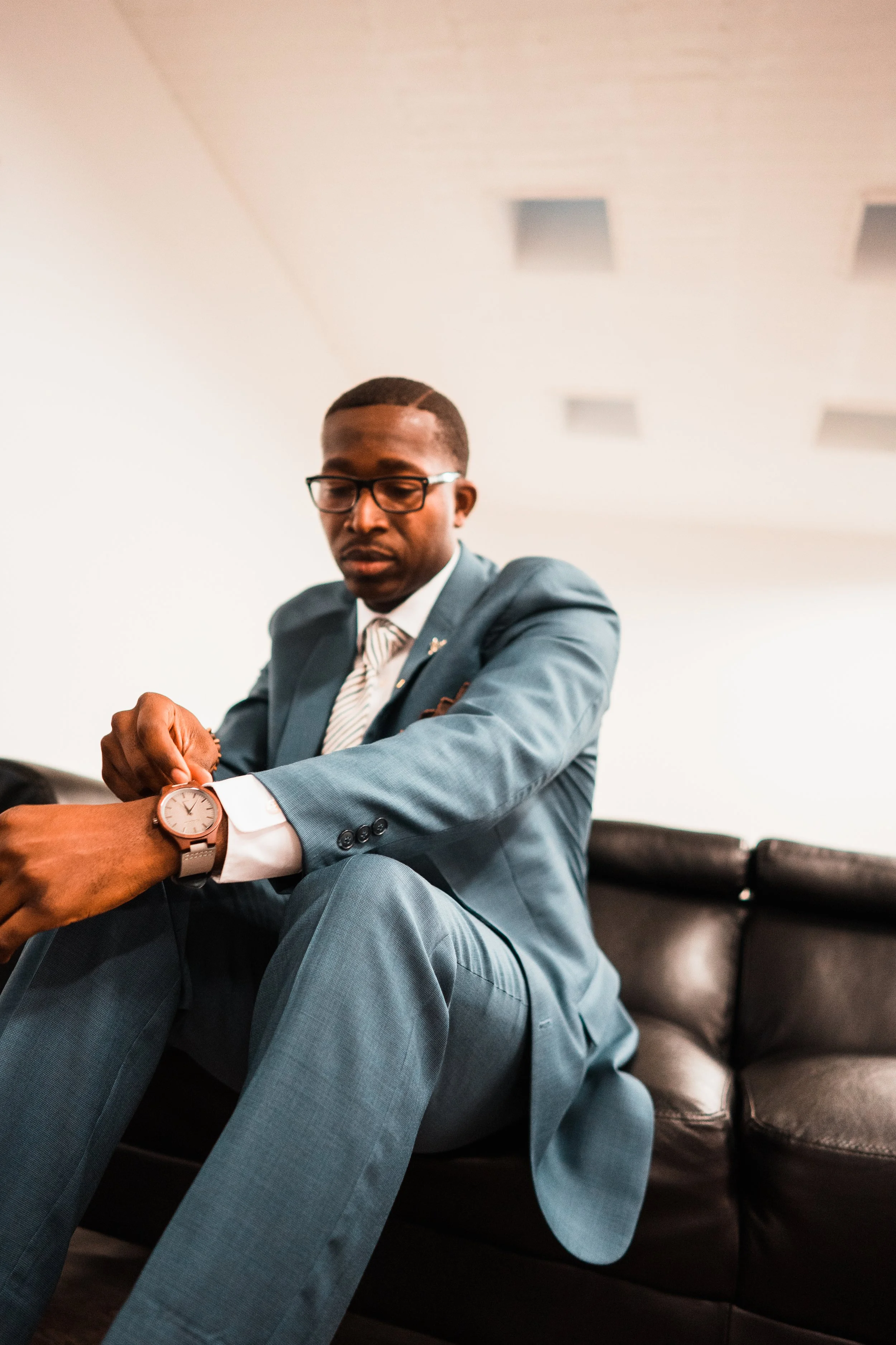 A man in a blue suit sitting on a black leather couch, adjusting his watch held by another person.