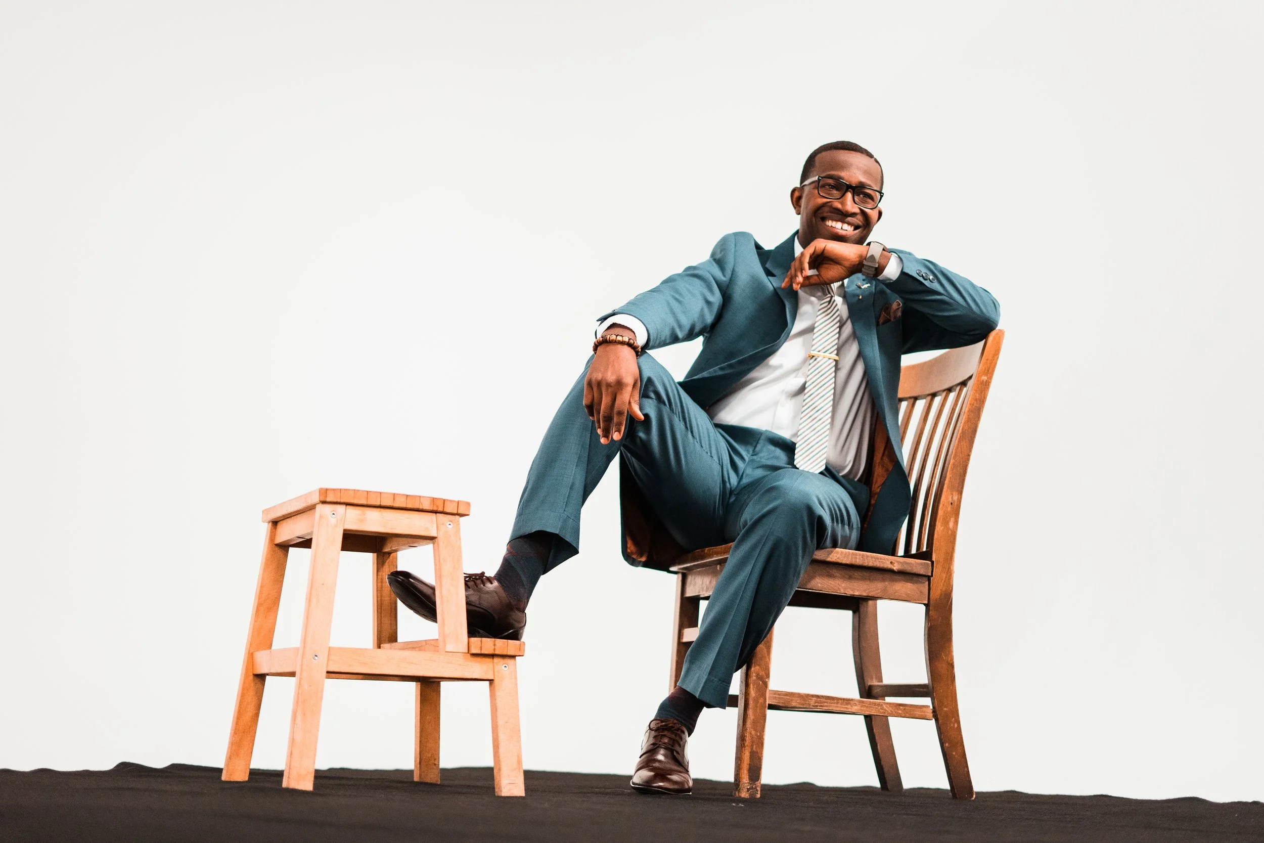 A smiling man in a teal suit, white shirt, and striped tie sitting on a wooden chair with one leg resting on a wooden stool, against a plain white background.